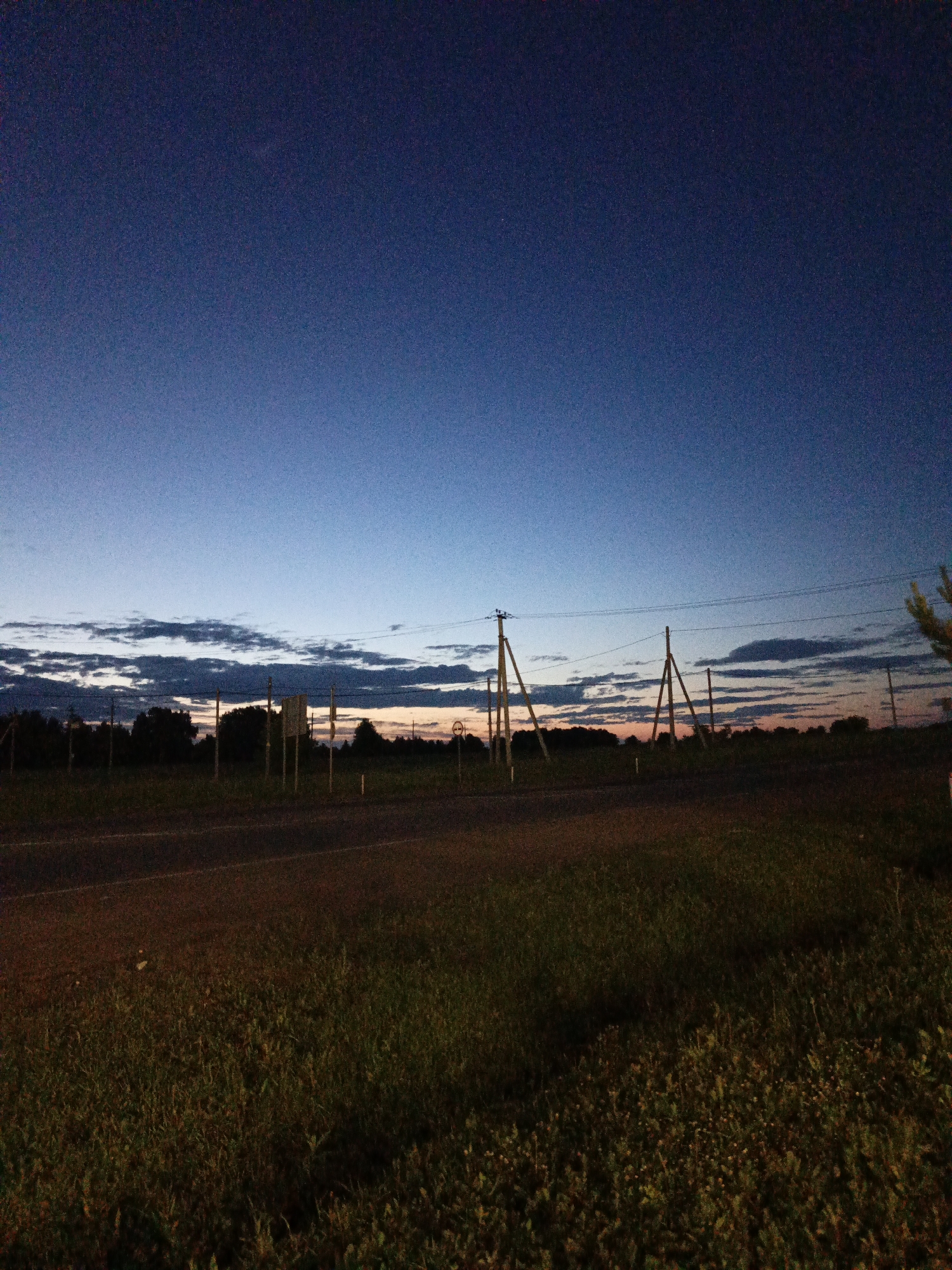 The image depicts a twilight scene at dusk in Kalachevo, Russia. A dark asphalt road occupies the lower third of the frame, bordered by a grassy field. Beyond the road, a line of dark trees forms a silhouette against the horizon. Several wooden utility poles with power lines are visible, some supporting what appear to be road signs. The sky is a gradient of deep blue at the top, transitioning to lighter blues and streaks of orange and pink near the horizon, indicating the sun has recently set. Scattered, dark clouds are present across the lower part of the sky. The overall environment suggests a rural or semi-rural roadside setting. There are no people or animals visible in the image.