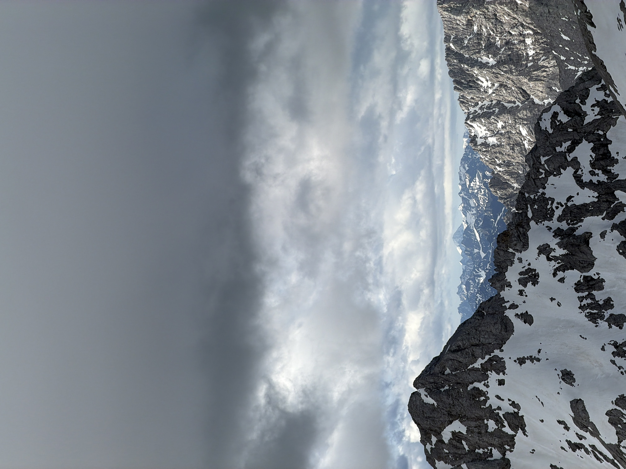 The image depicts a mountainous landscape with snow-capped peaks under a dramatic, cloudy sky. The foreground features a dark, rocky mountain slope partially covered in snow. Behind it, a series of rugged, snow-dusted mountain ranges extend into the distance. The sky is dominated by heavy, grey clouds, with patches of lighter clouds allowing some diffused light to filter through. The scene appears to be a natural, outdoor environment. The image does not contain any people, specific objects or actions that would indicate a city location.
