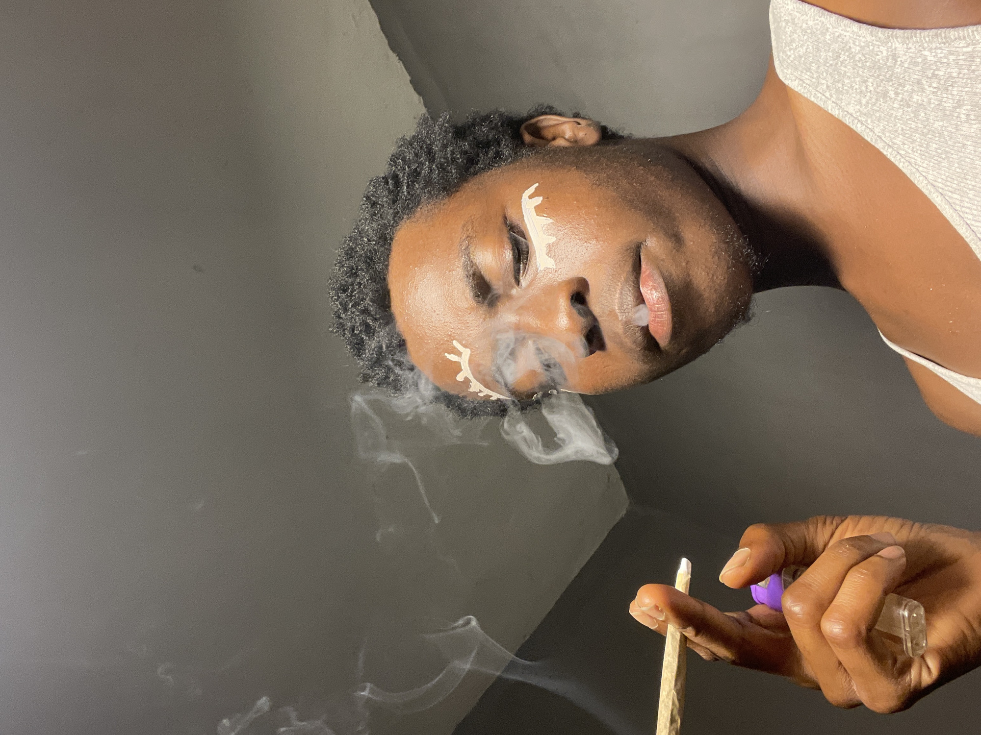 The image shows a close-up of a young Black person with dark, curly hair, wearing a grey tank top. They are holding a rolled cigarette or joint between their index and middle fingers, with a small purple object, possibly a lighter, held by their thumb. Smoke is emanating from the cigarette and around their face. The person has white paint or makeup applied around their eyes in a decorative pattern resembling eyelashes or tears. The background is a plain, dark grey wall. The scene is lit by a single light source, creating shadows. The image is a portrait, with the focus on the person's face and the act of smoking. The location is identified as Nkpor, Nigeria.
