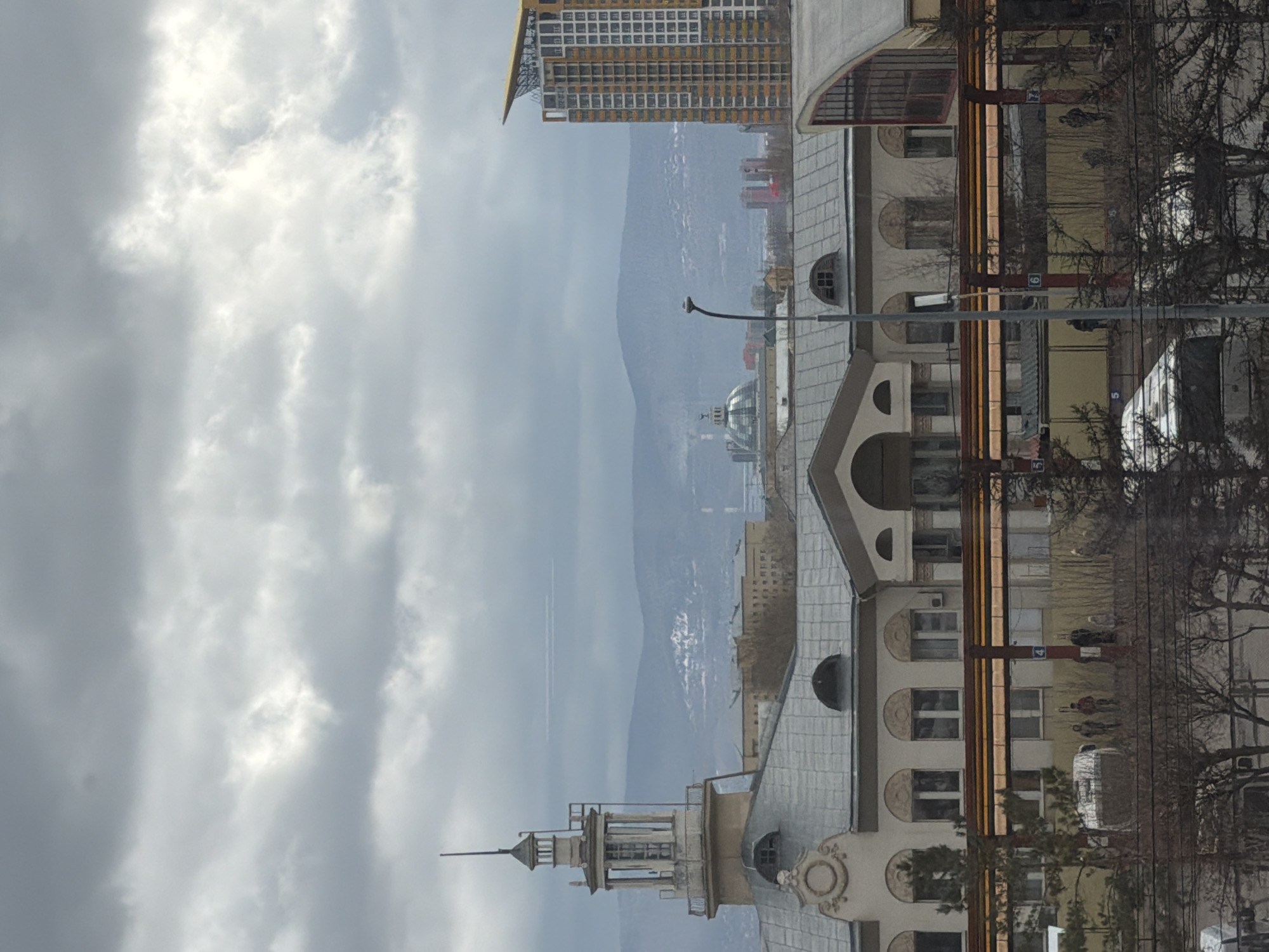 The image presents a cityscape in Krasnoyarsk, Russia, viewed on an overcast day. In the foreground, an ornate building with a prominent tower or spire featuring arched windows and a grey roof dominates the left side. Adjacent to it, another building with a similar architectural style and a grey roof is visible. At street level, bare trees suggest a winter or early spring season. Several vertical poles are present, one displaying the number "4" and another "5", likely indicative of a public transport stop. A few indistinct human figures are discernible near these poles, and a white vehicle, possibly a bus, is partially obscured by the trees.
The middle ground features a distinct, modern high-rise building with a yellow or gold upper section and numerous square windows. Further in the distance, a building with a glass dome can be seen, along with a street lamp. The background is dominated by large, undulating mountains, which appear partially covered in snow or frost, consistent with a cold climate. A faint airplane contrail is visible in the cloudy sky above the mountain range. The sky is entirely overcast, with areas of brighter, diffused light. The scene captures a typical daytime urban setting, devoid of any specific event beyond the ordinary flow of city life.