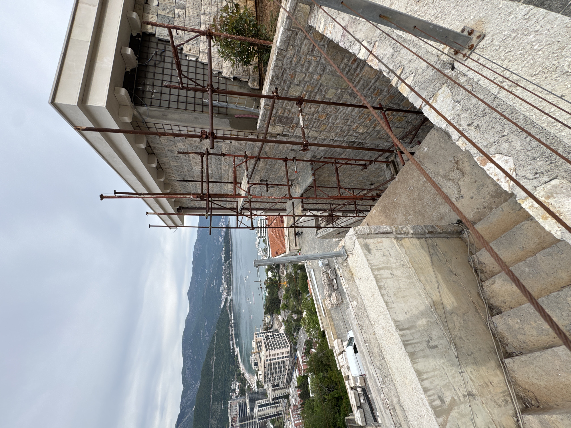 The image shows a partial view from a high vantage point in the city of Budva, Montenegro. In the foreground, stone steps and scaffolding are visible, suggesting ongoing construction or renovation work on a building. Beyond the scaffolding, a cityscape is apparent, including buildings, trees, and streets. A body of water, likely a bay or part of the Adriatic Sea, stretches into the distance, bordered by mountainous terrain. The sky is overcast, with light cloud cover.