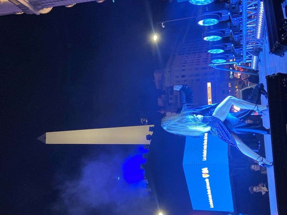 At night, a performer in a sparkling dress strikes a pose on stage, spotlighted by bright blue lights. The Obelisco stands tall in the background, partially obscured by haze. A screen displays text in Spanish. Stage lighting rigs are visible above the performer. A multi-story building can be seen behind the Obelisco. Some spectators are visible at the edge of the stage.