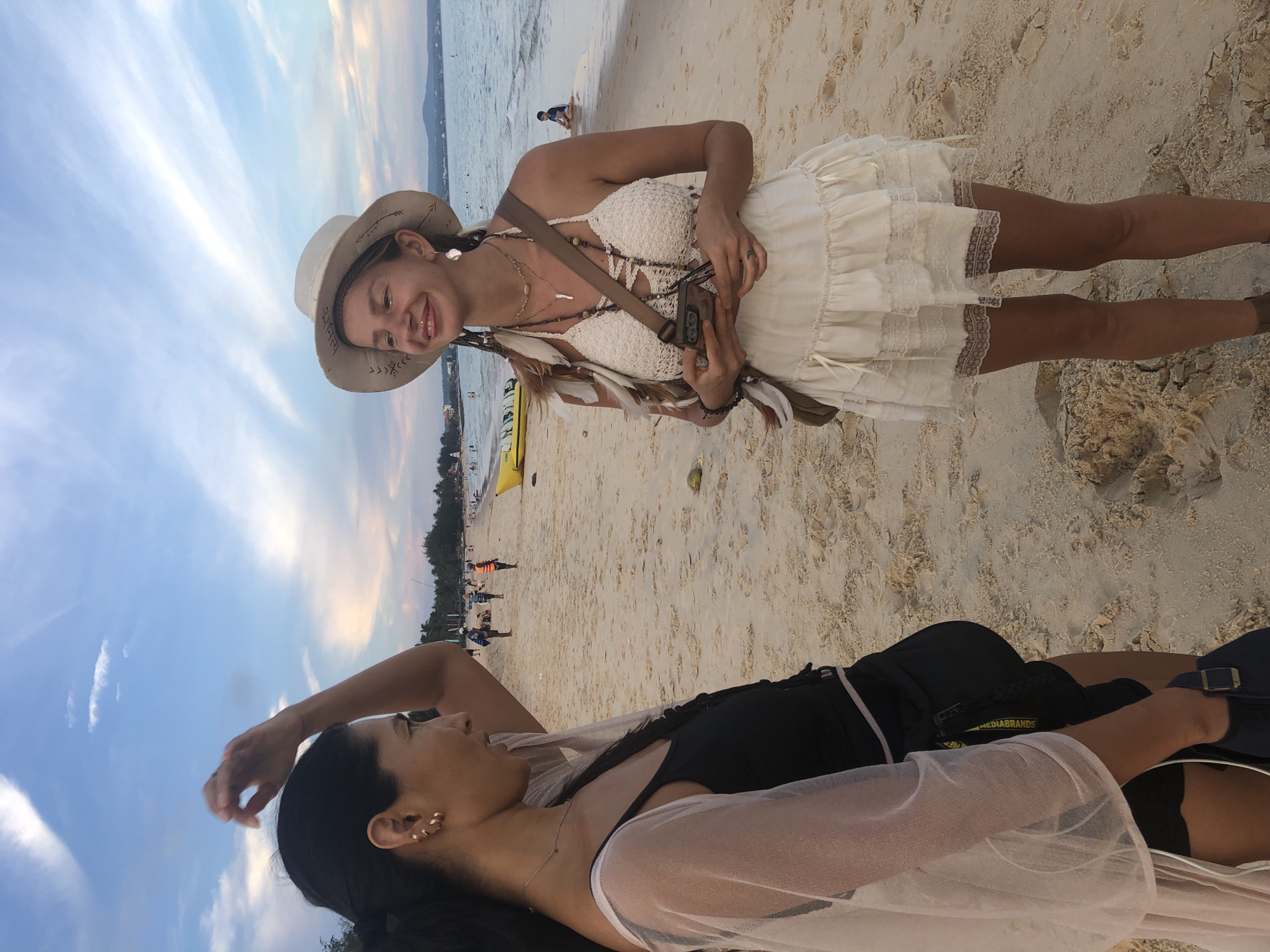 This image shows two women on a beach in Mũi Né, Vietnam. The woman on the left is wearing a black tank top and a sheer pink cover-up. She has her hair pulled back and is wearing hoop earrings. The woman on the right is wearing a white, bohemian-style dress and a wide-brimmed hat. She has feathers woven into her hair and is wearing multiple necklaces and earrings. Both women are looking at each other and smiling. The background shows a sandy beach with people in the distance, some in the water, and trees and buildings along the coastline. The sky is cloudy with hints of pink and orange from a sunset.