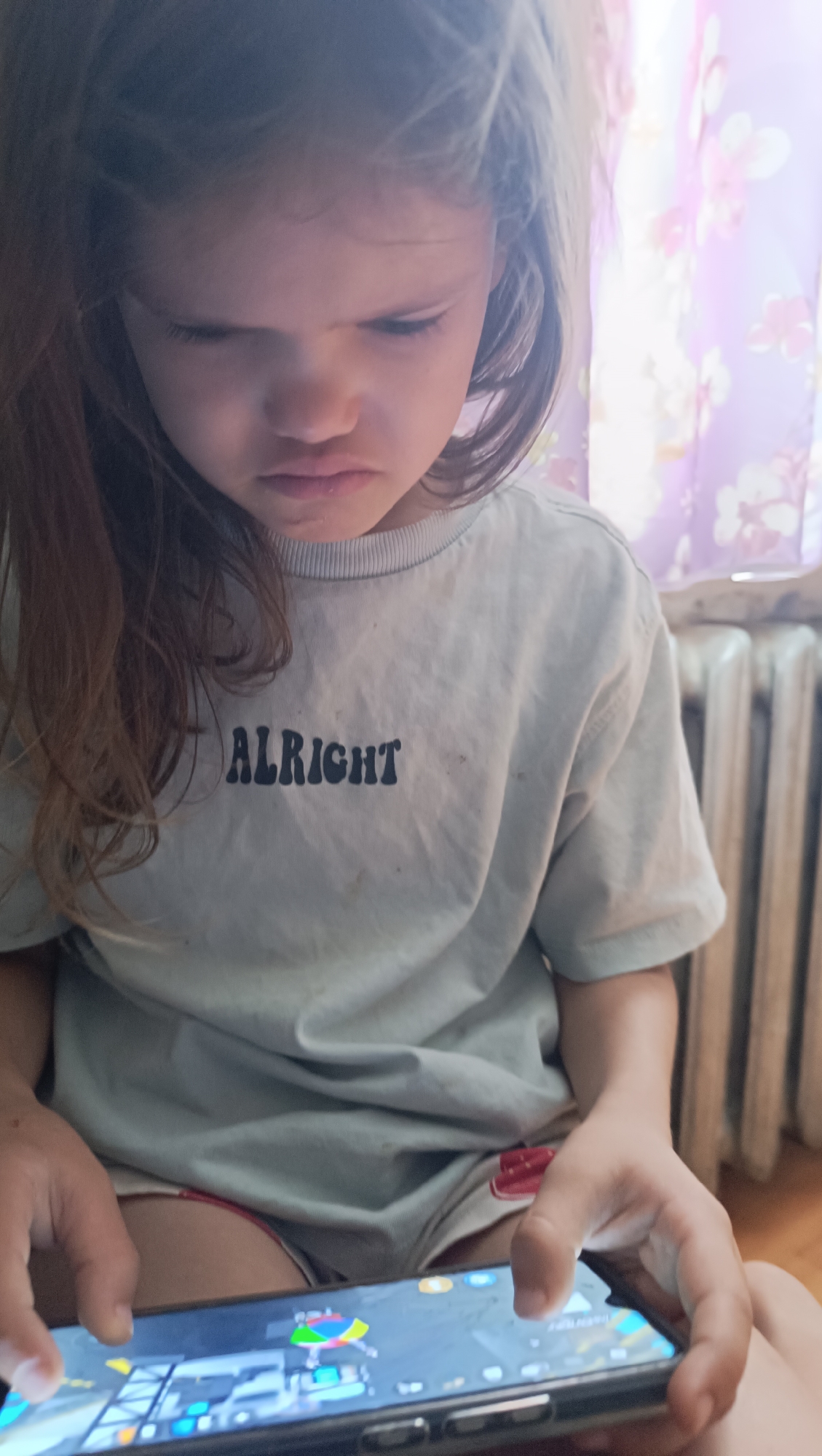 A young girl with long brown hair is sitting and playing a game on a smartphone. She is wearing a light blue t-shirt with the word "ALRIGHT" printed in black letters. Her facial expression appears serious or perhaps frustrated, as she is looking down intently at the phone. Her hands are on the phone, actively engaged with the game. The setting seems to be indoors, with a radiator and a purple floral curtain visible in the background, suggesting a home environment. The lighting is soft, possibly indicating daytime or a room with natural light coming from a window. The image captures a moment of focused engagement with a digital activity.