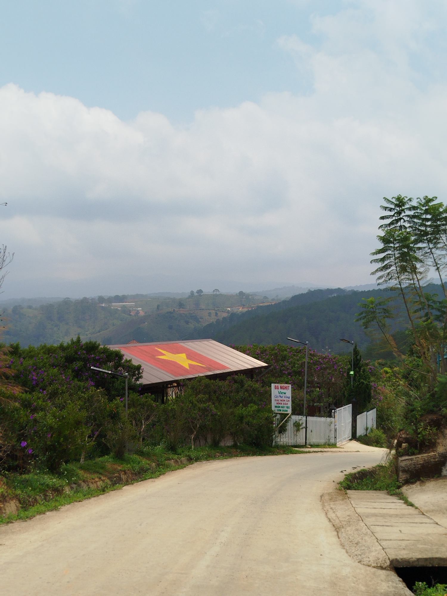 The image depicts a rural scene in Đa Nghịt, Vietnam. In the foreground is a dirt road curving around a corner. Adjacent to the road is a concrete drainage ditch. Lush greenery, including bushes and small trees, lines the roadside. In the midground, a building with a roof painted with the flag of Vietnam is visible. A sign in Vietnamese that says "ĐA NGHỊT View Cà Phê Vòng CÁC MÓN ĂN BÌNH DÂN NHẬN ĐẶT TIỆC CƠM ĐOÀN" is placed in front of a white fence. Hills covered with forests and possible cultivated areas are in the background under a cloudy sky.