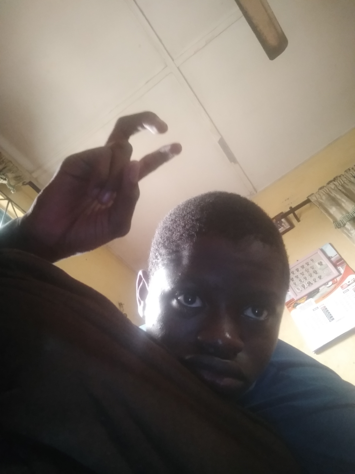 A low-angle indoor shot shows a dark-skinned person with close-cropped hair. The person is holding up their hand, making a "peace" or "victory" sign. The room has a ceiling fan and what appears to be a calendar on the wall behind the person. The image's composition is off-center. The background includes what appears to be a window with curtains and potentially other furnishings. The location is indiscernible as Shangisha, Nigeria.