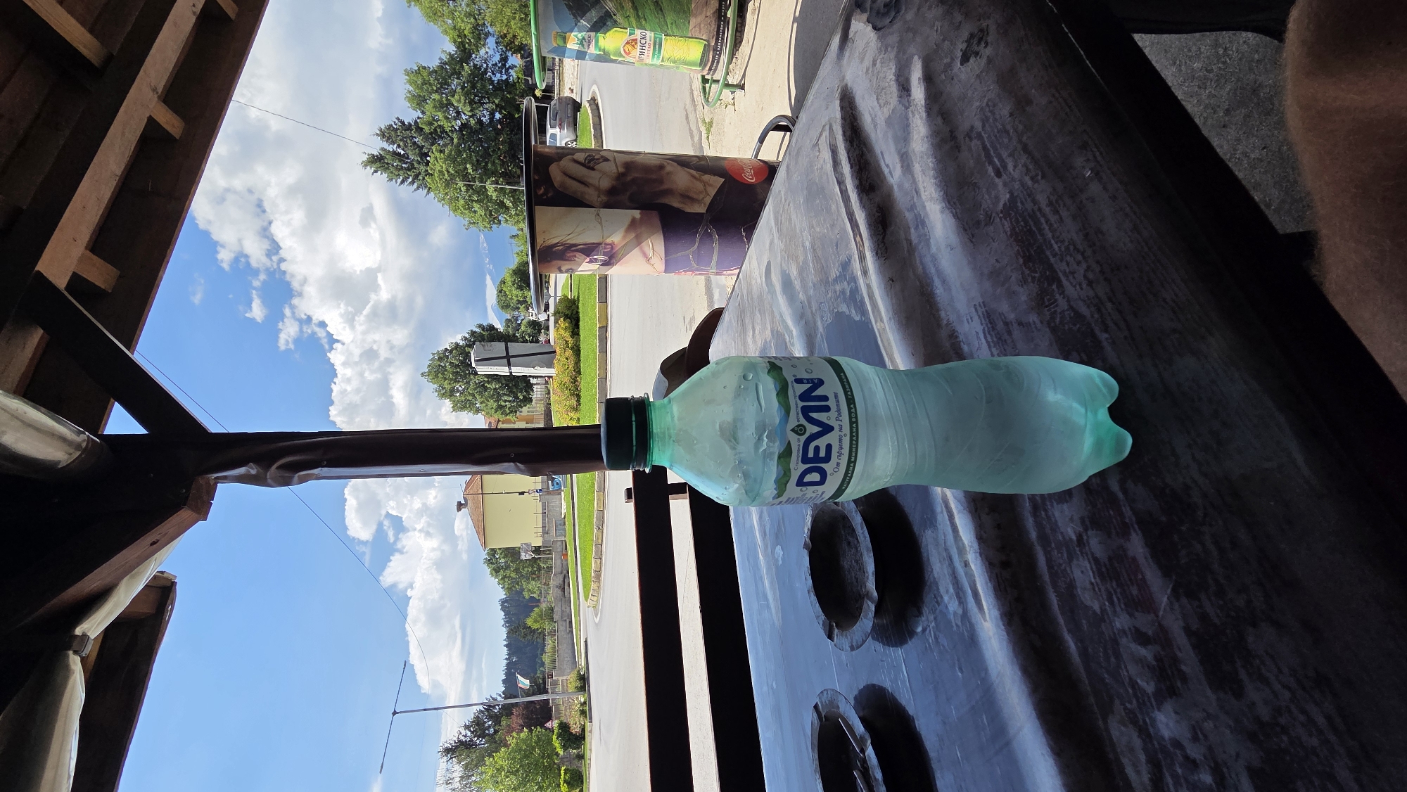 A bottle of Devin mineral water sits on a wooden table in front of two ashtrays. Behind the table, a street scene unfolds with a roundabout, buildings, trees, and a bright blue sky with white clouds. To the right, a cylindrical display featuring an image of a woman and a Coca-Cola advertisement is visible, along with another advertisement for beer. The overall impression is of an outdoor seating area at a cafe or restaurant.