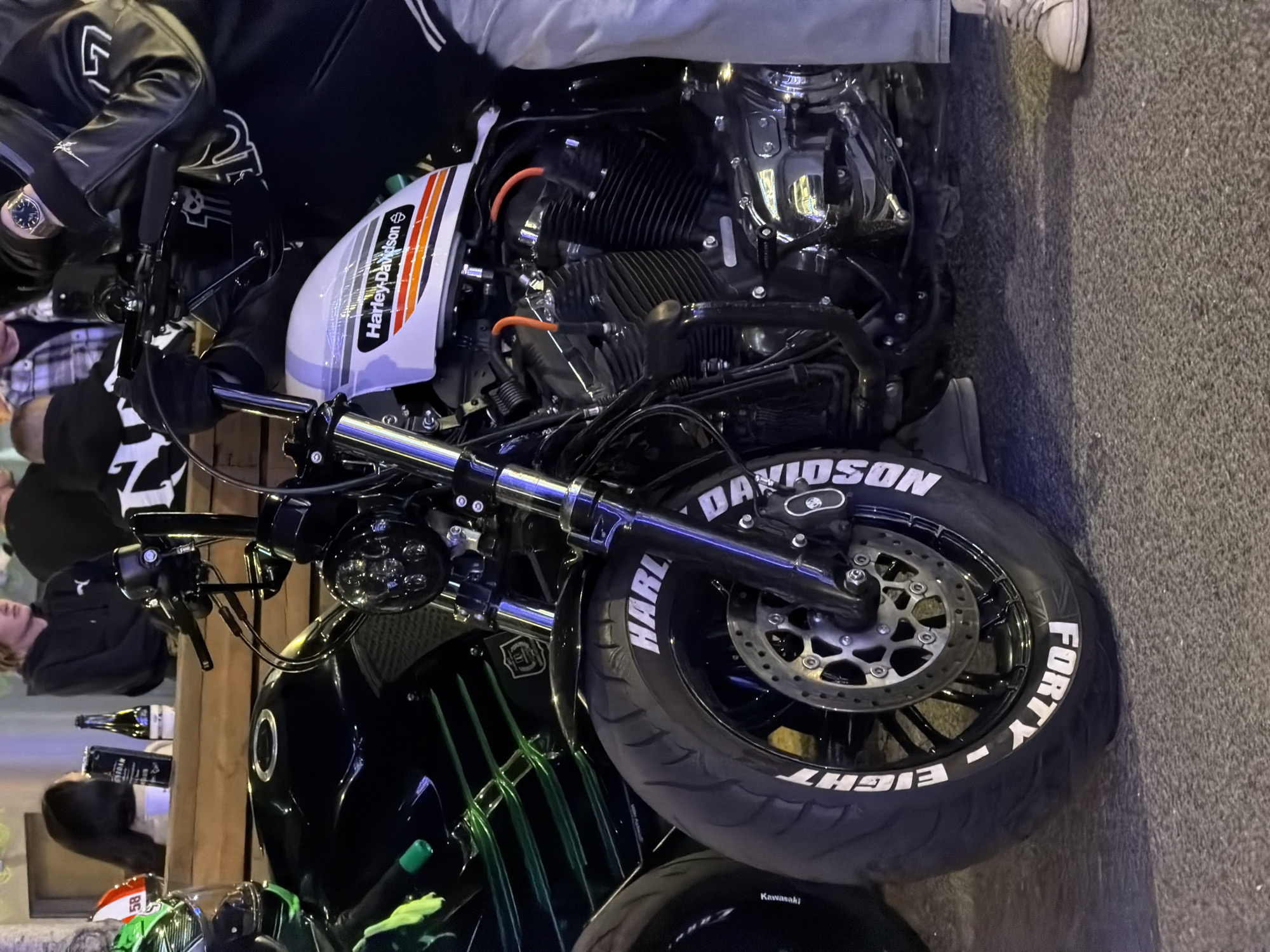 The image shows a close-up of a Harley Davidson Forty-Eight motorcycle parked in an urban environment, likely in a European city, possibly Moscow, Russia. The motorcycle has a white fuel tank with the "Harley-Davidson" logo and orange and grey stripes. The front tire has "Harley-Davidson" and "Forty-Eight" written on it in white lettering. Parts of other motorcycles, including what appears to be a Kawasaki, are visible nearby, suggesting a gathering of motorcycles. People are visible in the background, standing and socializing near a wooden structure or bar. Bottles and glasses are on the bar. The pavement is asphalt. The time of day appears to be evening or night, as the scene is illuminated by artificial lighting.