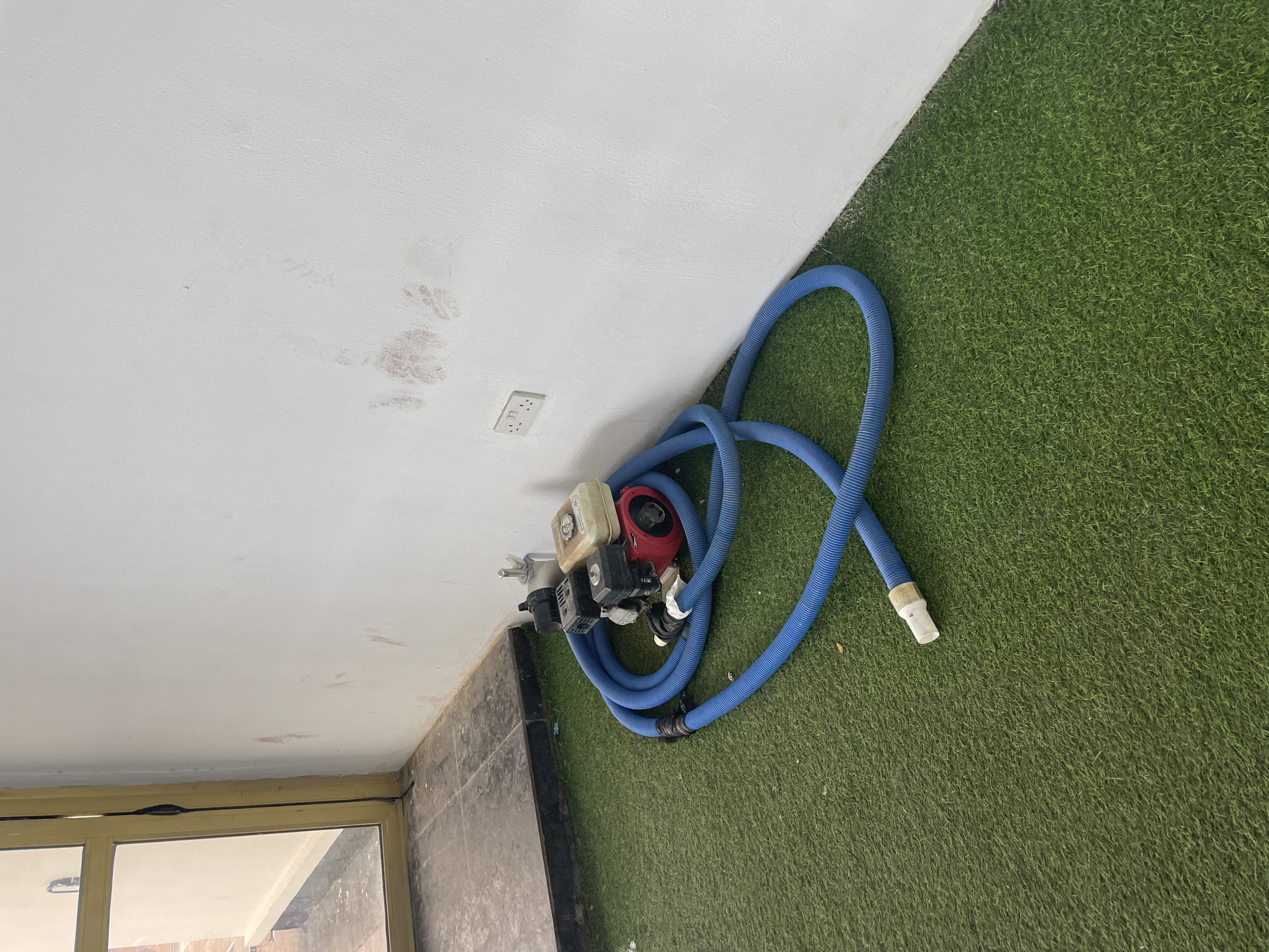 The image depicts a corner of an indoor space with artificial turf flooring. A red and beige engine with a black motor is placed on the turf, coiled around it is a long blue flexible hose. To the left of the engine, there is a metal fitting. The wall behind the engine is white and shows some reddish-brown smudges. There is a white electrical outlet on the wall. To the far left, a glass door with a yellow frame is partially visible, leading to an outdoor area with tiled flooring. The scene is devoid of any individuals. The image does not provide specific information about the location in Jauro Yinu, Nigeria.