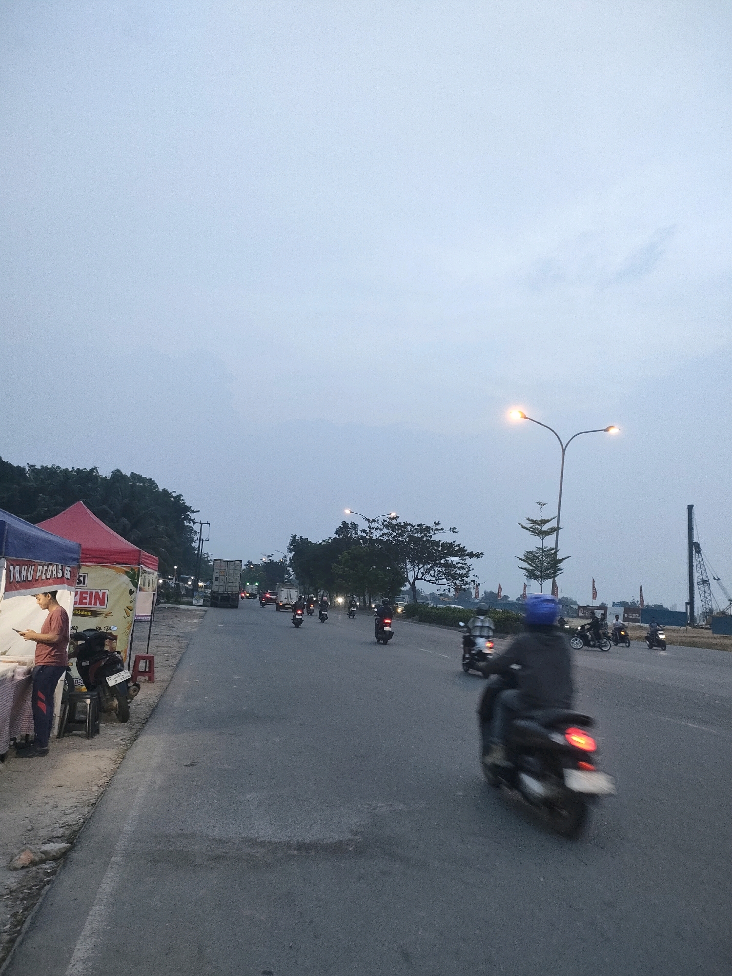 The image depicts a street scene in Batam, Indonesia, likely near a busy thoroughfare at dusk. In the foreground, a person stands by a food stall, which is covered by a red and blue awning. The stall displays text that is partially visible, including "RHU PEDAS" and "LEIN." A motorcycle is parked beside the stall.
The street itself is paved and has a white line marking its edge. Several motorcycles are in motion on the road. One motorcycle in the mid-ground, ridden by a person wearing a blue helmet, is moving away from the viewer. Further down the road, more motorcycles and a truck are visible. On the right side of the road, a large street lamp illuminates the scene, and construction equipment, including a crane, can be seen in the background. Trees line the left side of the road. The sky is overcast with a pale blue hue.
