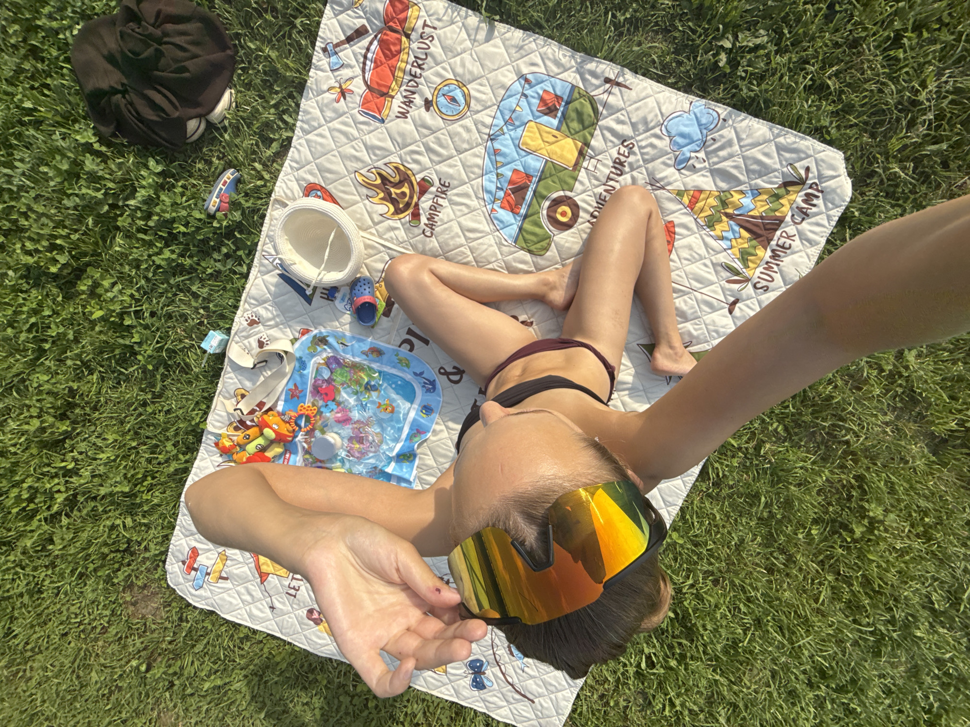 This image shows a person lying on a picnic blanket spread on green grass. The person is wearing a bikini and large, reflective sunglasses. The picnic blanket has a pattern featuring camping-themed illustrations like a camper van, a tent, and the words "WANDERLUST," "CAMPFIRE," "ADVENTURES," and "SUMMER CAMP." There are also several objects scattered around the blanket, including a black bag with shoes inside, a child's sandal, a white sun hat, a bucket, and a small inflatable water play mat with toys. The scene appears to be outdoors in a grassy area, likely a park or backyard, under natural sunlight, suggesting it is daytime. The person's pose and the presence of the water play mat suggest a relaxed, leisurely activity, possibly a summer outing or picnic. The weather seems pleasant, given the clear sunlight. The location context provided is Podshivalovo, Russia.