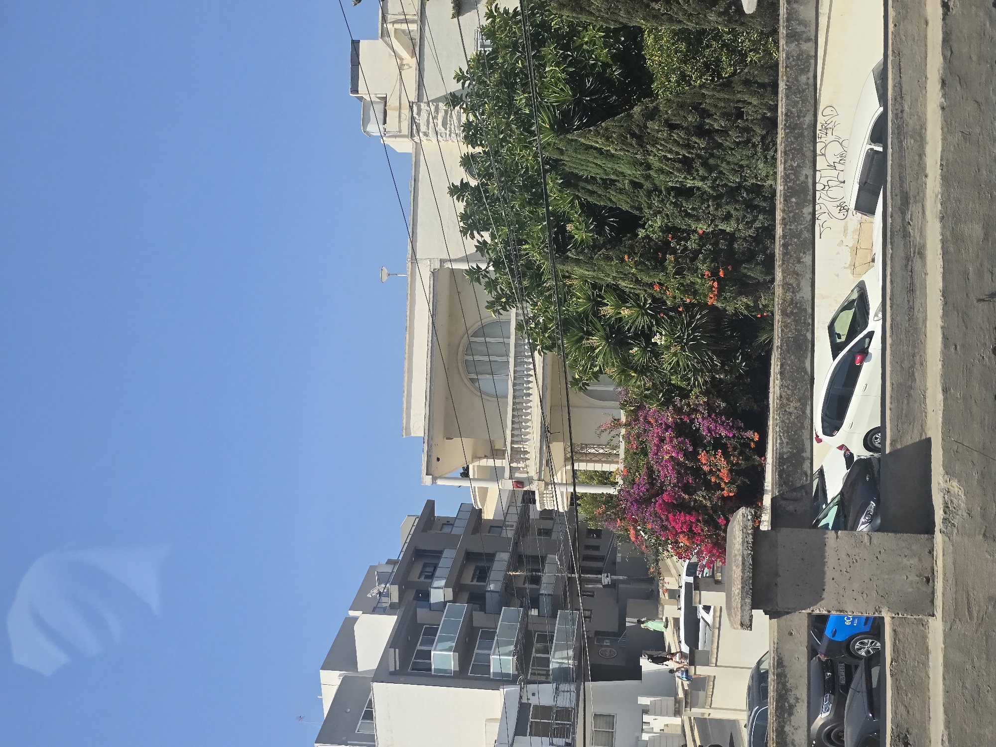 In Swieqi, Malta, the image captures a street scene under a clear blue sky, with several modern apartment buildings and a detached villa. On the left, a multi-story apartment building features a grey facade and balconies with glass railings. To its right, a lighter-colored villa with classical architectural elements, including columns and a prominent arched window, is visible. A dense arrangement of trees and flowering bushes, displaying vibrant pink and orange blooms, partially obscures the lower portions of the buildings and the street. Several cars are parked along the street, including a black SUV, a blue and black electric vehicle, a white hatchback, and other vehicles partially visible. A concrete wall runs along the foreground, separating the viewer's perspective from the street level. The overall scene depicts a sunny day in a residential area with a mix of modern and traditional architecture.