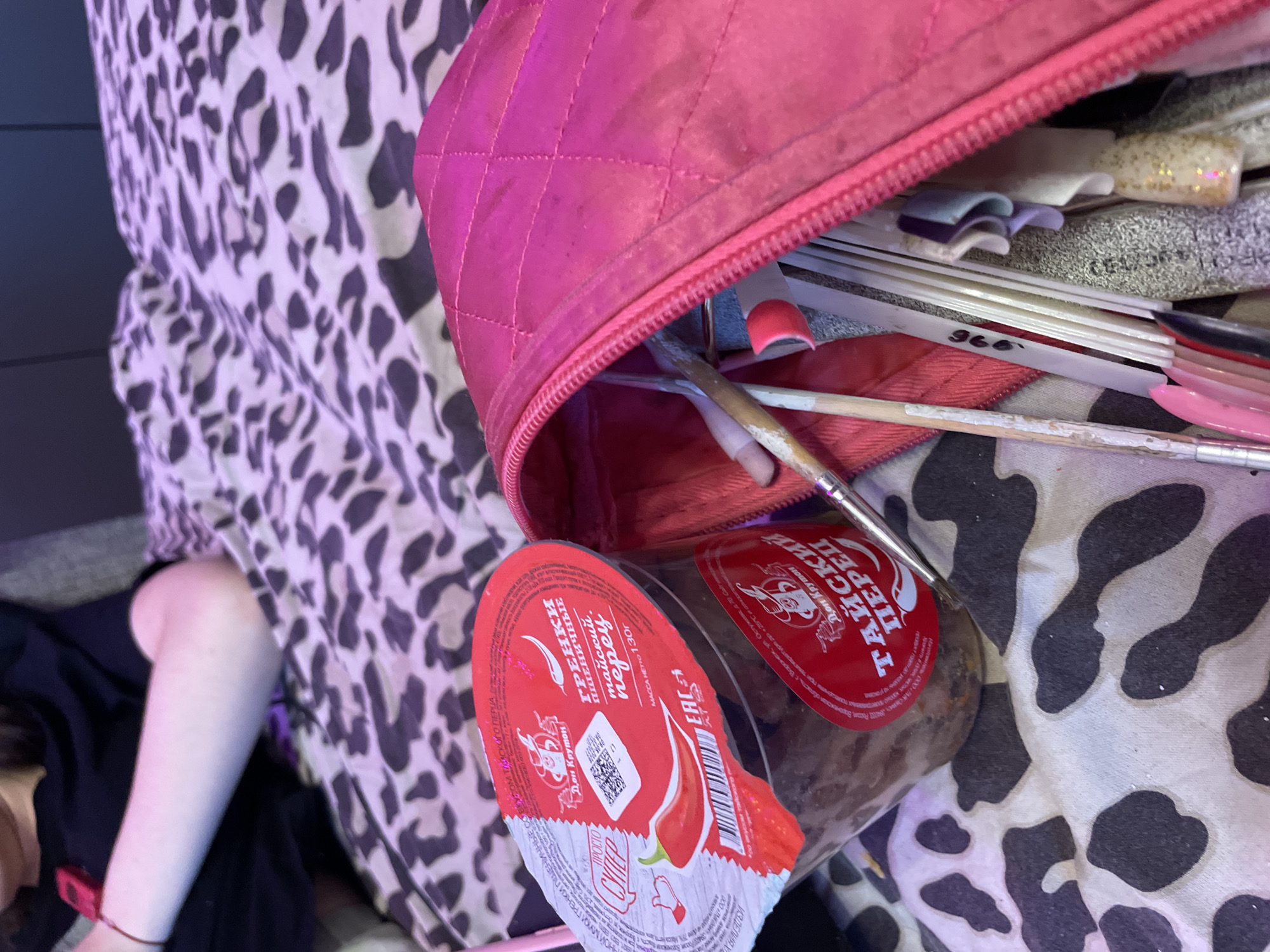 The image shows a close-up view of a person's arm and a pink quilted bag filled with nail art supplies and a container of food. The person's arm is resting on a patterned surface, possibly a bed or sofa, with a black shirt visible on the upper body. The pink bag is partially open, revealing various tools, brushes, and nail art items like artificial nails with glitter. A red-lidded plastic container with Cyrillic labeling, appearing to contain food items, is placed next to the bag. The overall environment suggests a personal space, likely indoors, with a focus on creative activities or a break from them. The location is Ladozhskoe Ozero, Russia.