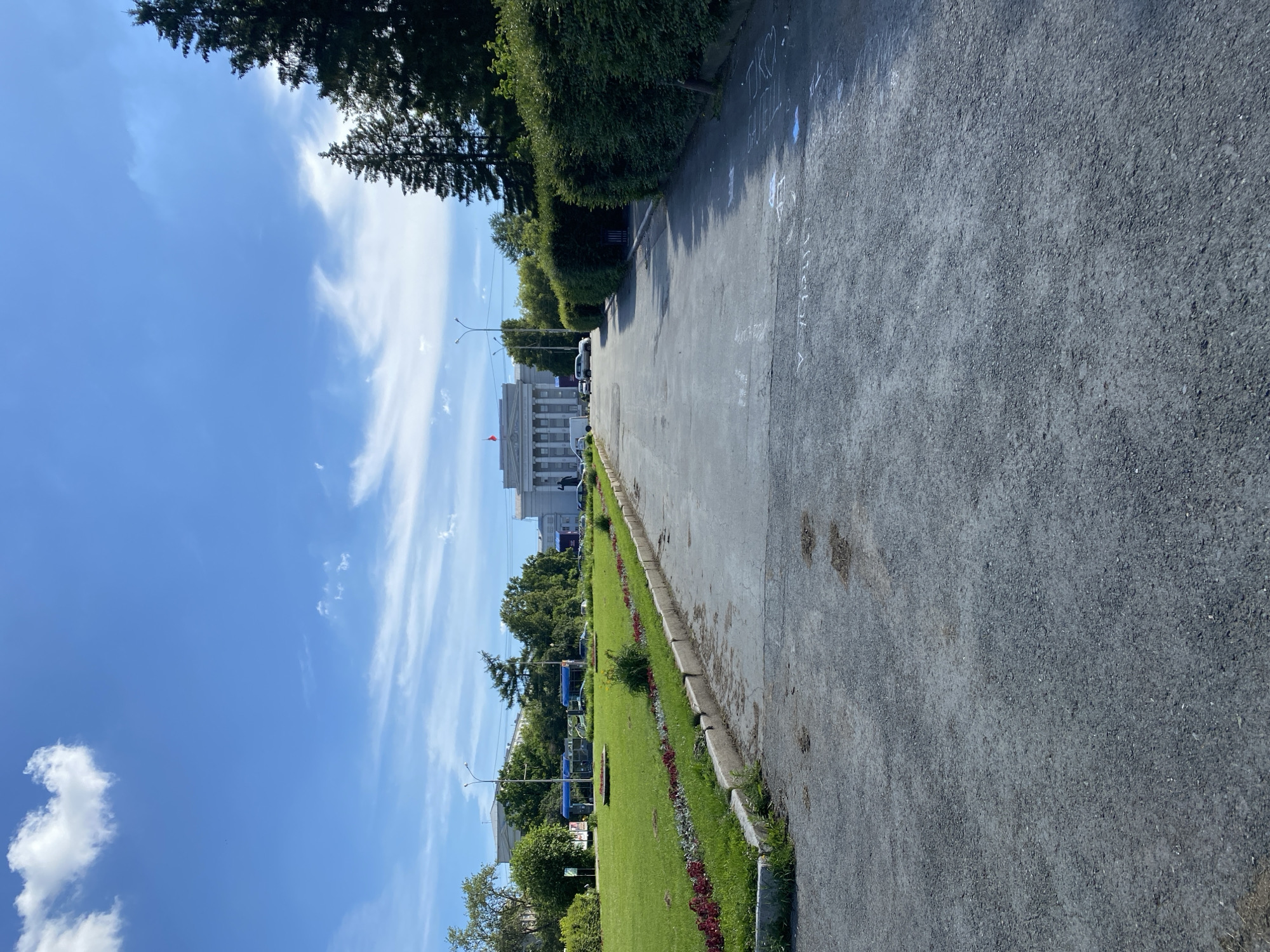 The image shows a wide paved pathway leading towards a grand white building with columns. The building, identified as the Yekaterinburg City Duma, is situated in a park-like setting. On the left side of the pathway, there is a neatly manicured lawn with flowerbeds. Further in the background, lush green trees and smaller buildings are visible. The sky above is a clear blue with scattered white clouds, suggesting fair weather. A few cars are parked in front of the main building. The overall scene depicts a public space in Yekaterinburg, Russia, on a bright day.