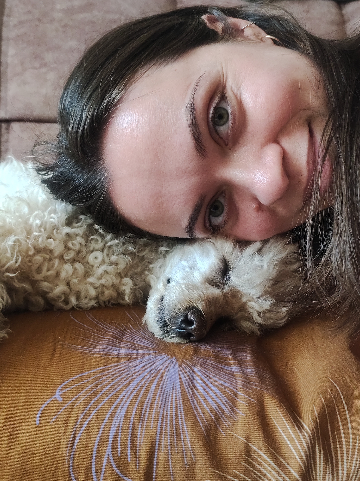 A close-up, slightly angled overhead shot shows a woman with long, dark hair and green eyes lying down with a small, fluffy white dog. The woman is smiling faintly. Her head rests on the dog's body. The dog, possibly a poodle mix, has its eyes closed and appears to be sleeping. Both are lying on a mustard-yellow pillow or surface with a faded, stylized flower design. The location, Chaikovsky, Russia, is not visually discernible from the image alone.