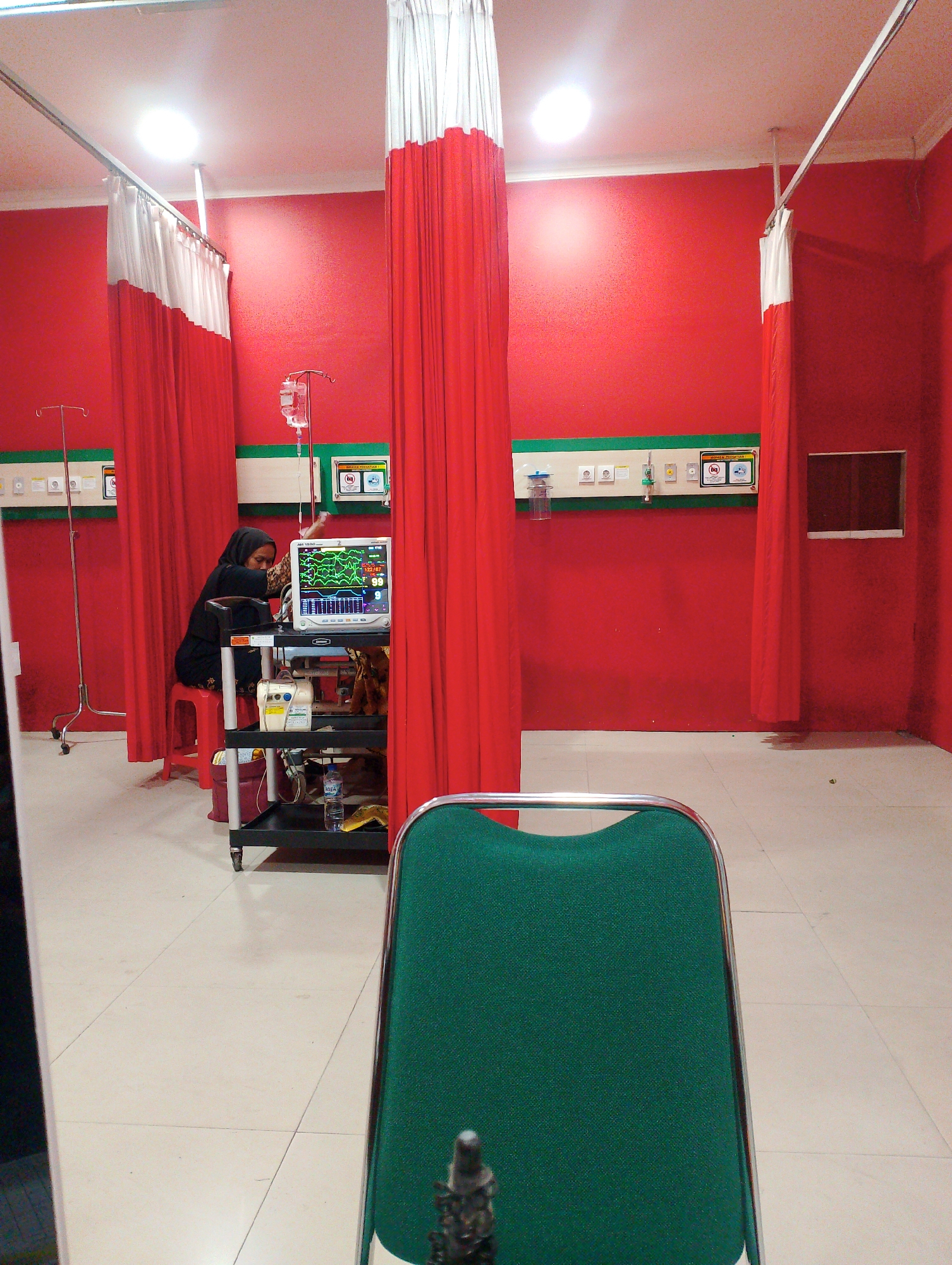 The image depicts an indoor scene within a medical facility, possibly an emergency room or a treatment area. The room is painted a vibrant red, with red and white curtains that appear to be partition dividers. The floor is tiled in a neutral, light color.
In the mid-ground, to the left, a woman wearing a black hijab and dark clothing sits on a red stool. She is positioned next to a medical cart which holds a vital signs monitor displaying a patient's readings, an IV bag hanging above, and other medical equipment. To the far left, a metal IV stand is visible.
The wall features a green panel with various medical gas outlets and electrical sockets. On the right side of the image, a dark rectangular window or opening is present.
In the foreground, the back of a dark green upholstered chair with a metal frame is prominently displayed, suggesting the perspective of someone observing the scene from a seated position.
The overall impression is one of a functional, albeit somewhat stark, medical environment, likely in Sidoarjo, Indonesia, given the context provided. The presence of medical monitoring equipment and the general setup suggest a patient care area.