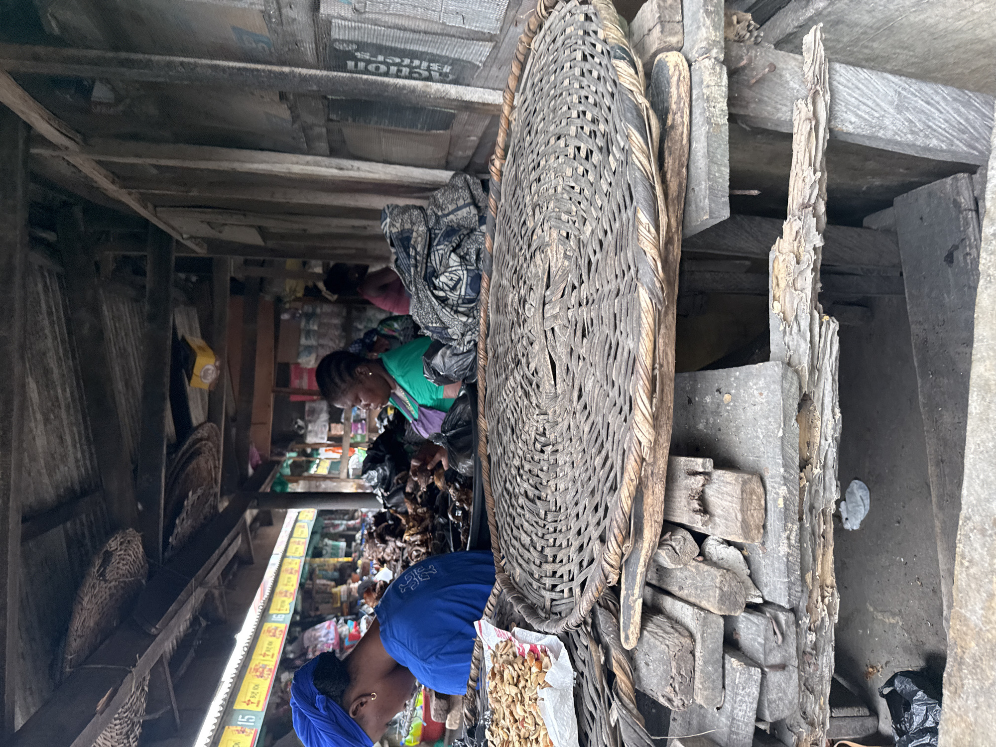 The image depicts an outdoor market stall in Lagos, Nigeria, likely a food market given the presence of dried goods and various food items. In the foreground, a large, woven circular tray made of natural fibers sits atop a stack of weathered wooden planks and flat stones. To the left of the tray, a small pile of dried, nut-like food items is displayed on a white paper.
Behind the tray, two women are partially visible. The woman on the left, wearing a blue t-shirt and a blue headwrap, is facing away from the camera, seemingly attending to her stall. The woman on the right, dressed in a green top, is looking down and appears to be interacting with items within her stall. Other figures are faintly visible in the background, suggesting a busy market environment.
The stall itself appears to be constructed from rough, weathered wood and corrugated metal roofing, typical of informal market structures in Lagos. Various items, including sacks of produce and what appear to be more woven baskets, are stored and displayed within the stall. The ground beneath the stall is dusty and uneven. The overall scene conveys a sense of daily commerce and the vibrant, often rustic, atmosphere of Lagos markets.