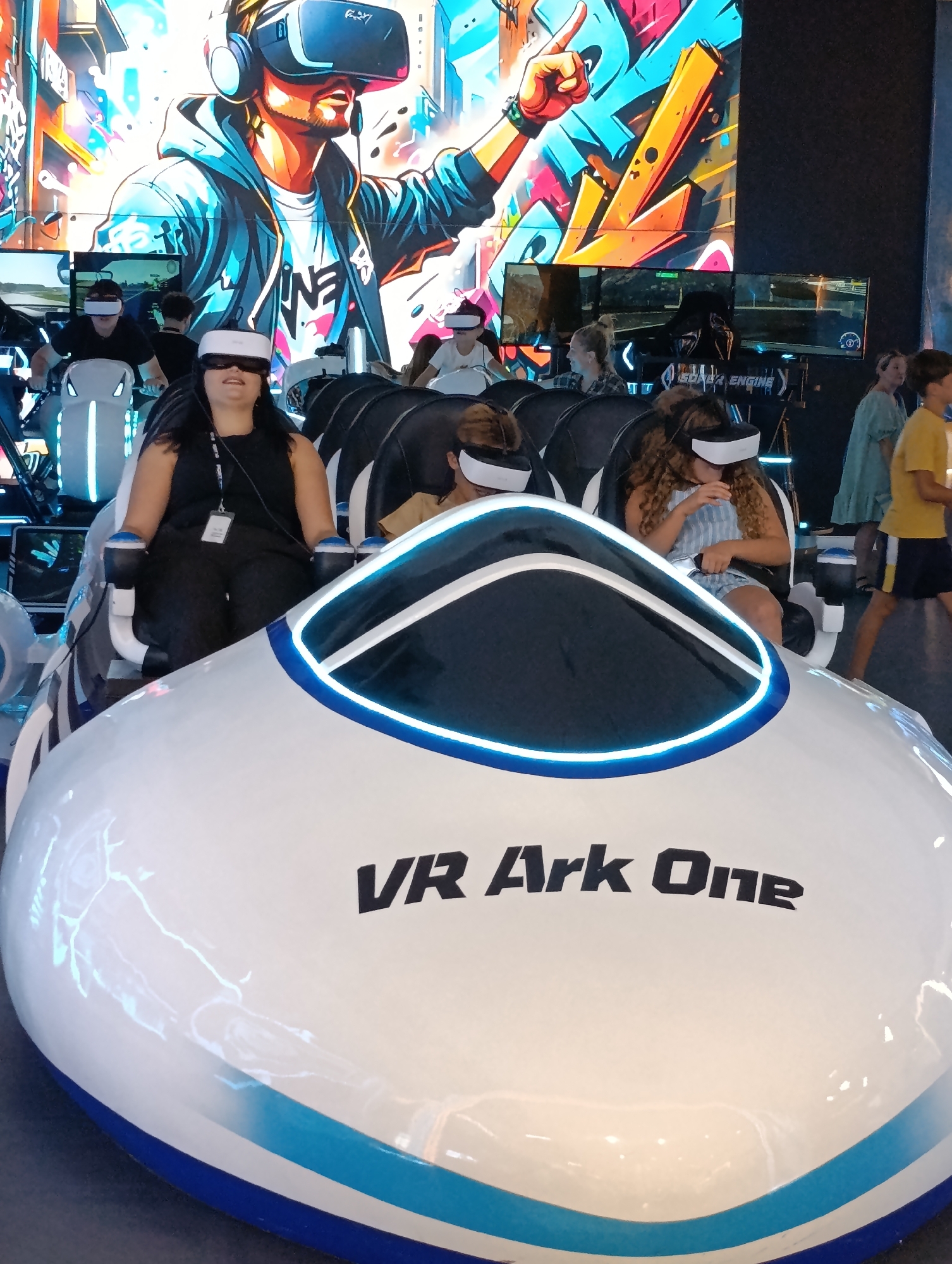 The image shows a VR Ark One attraction with several people wearing VR headsets seated in pods. In the foreground, a white pod with blue and black accents is visible, with the text "VR Ark One" printed on it. A woman in a black top and pants is seated in one of the pods, wearing a VR headset. Behind her, other individuals are also seated in pods, wearing VR headsets and appearing to be engaged in a virtual reality experience. In the background, large screens display graphics related to virtual reality. The overall environment appears to be an indoor entertainment venue. Without additional information or context, the specific location in Chișinău, Moldova cannot be determined from the image itself.