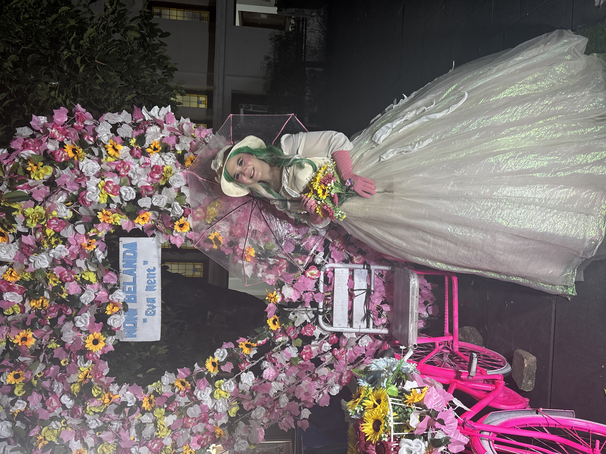 A full-shot, eye-level, outdoor nighttime photograph depicts a young woman dressed in a voluminous, light-colored, shimmering gown, complete with pink gloves and a straw hat, standing to the right of a large heart-shaped floral arrangement. The arrangement is composed of pink, white, and yellow artificial flowers and leaves, with a white sign reading "NON BELANDA" and "Eva Ment" hanging in the center. The woman is holding a bouquet of flowers and an open pink-rimmed umbrella. To the left of the floral arrangement is a bright pink bicycle with a basket full of flowers and a small metal chair. The background features a building with illuminated windows and lush green trees. The scene is likely set in the city of West Jakarta, Indonesia, given the context of the floral arrangement's inscription.