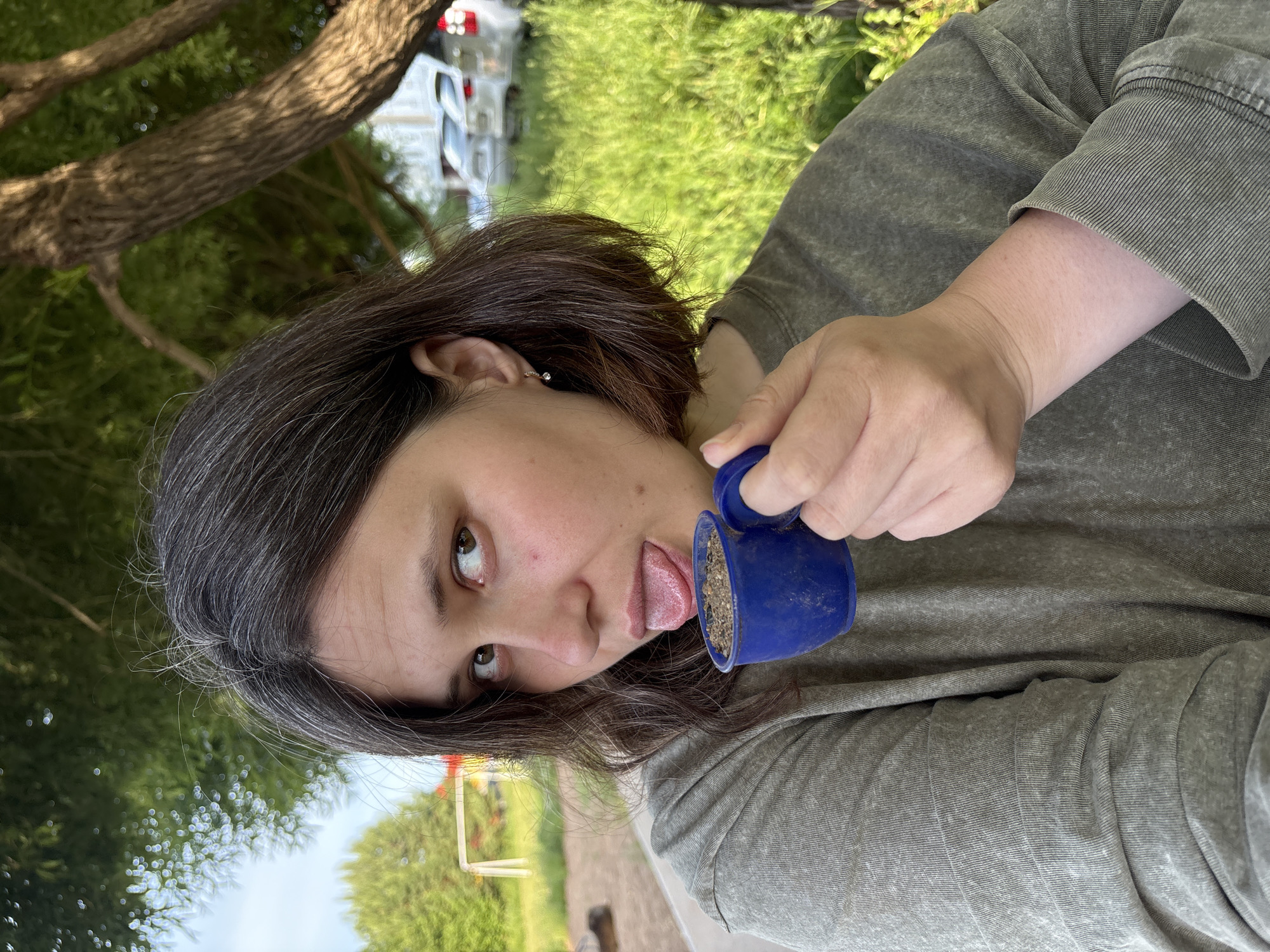 A playful outdoor portrait in Chaikovsky, Russia, features a person with dark, medium-length hair and fair skin. The subject, likely female, is shown from the chest up, looking directly at the camera with a mischievous expression, her head tilted, and her tongue playfully extended. She holds a small, well-used blue cup in her right hand, which contains what appears to be dirt, sand, or small pebbles, towards which her tongue is directed. She is wearing a comfortable-looking grey-green long-sleeved top. The background is a bright, sunlit natural setting with lush green trees and grass. Out-of-focus elements in the distance include what seem to be white vehicles and a light-colored structure, suggesting a park, yard, or recreational area. The image conveys a sense of humor and lightheartedness, captured during the daytime. No text is visible in the image.