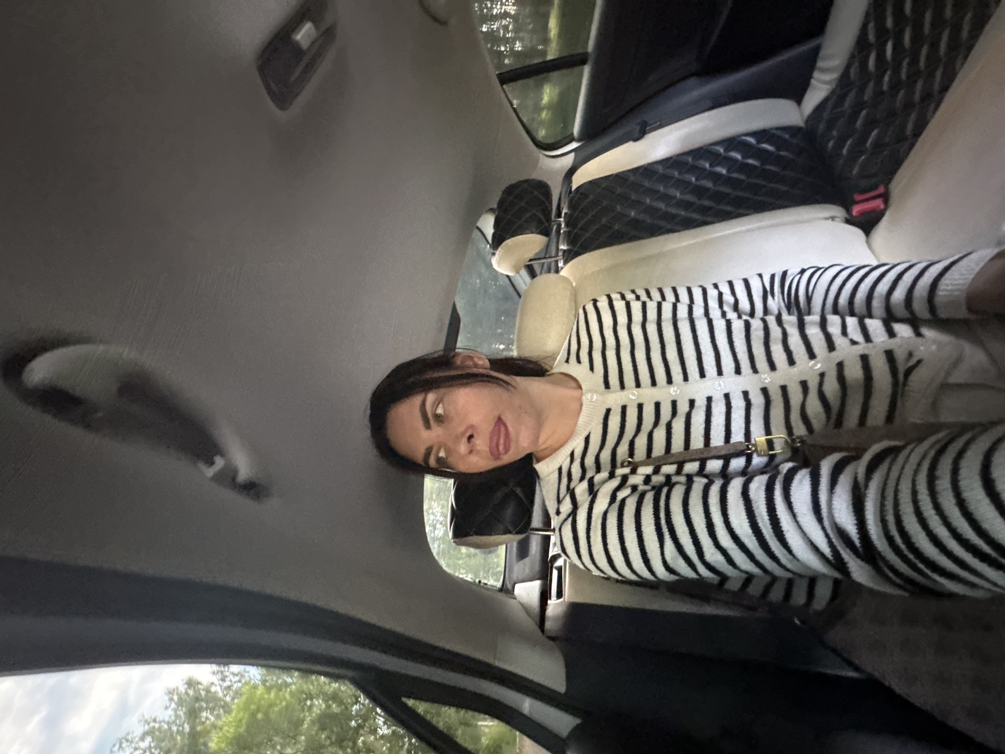 The image shows a woman sitting in the back seat of a car, looking out the window. She is wearing a black and white striped cardigan. The car interior is visible, including the seats, headrests, and ceiling. Outside the car, trees and a cloudy sky are visible. The location is specified as Chaikovsky, Russia. The woman has dark hair and is looking to her right with her tongue sticking out slightly. She is holding a purse on her lap. The seats have a quilted design.