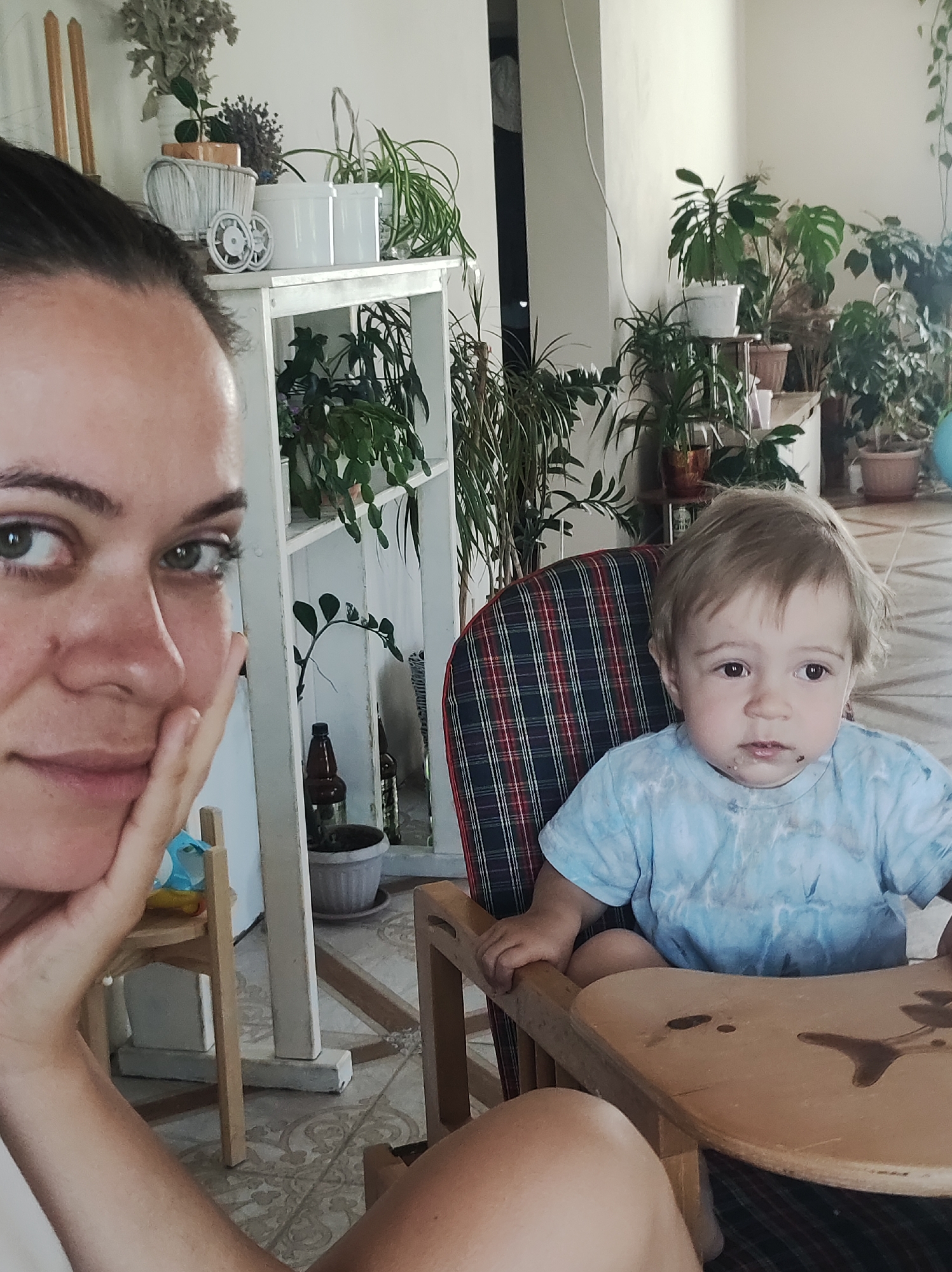 In the city of Fokino, Russia, a selfie captures a close-up of a woman's face on the left, with her hand resting on her chin. To her right, a young child sits in a high chair, wearing a blue tie-dye t-shirt. The child's face is slightly messy, suggesting recent eating. The background reveals an indoor environment filled with numerous potted plants on shelves and in various locations, creating a green and lively atmosphere. The flooring appears to be tiled.