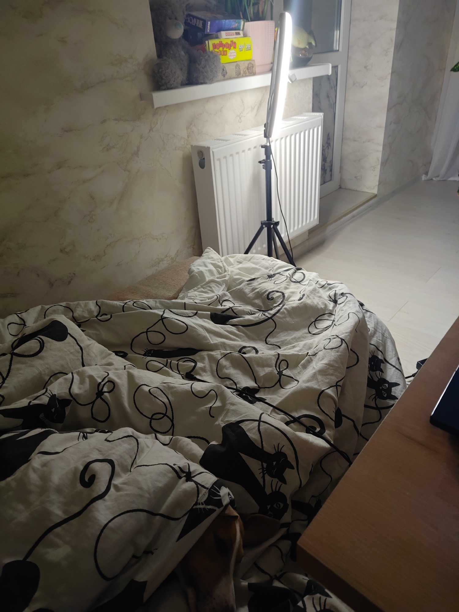 The image depicts an indoor scene, likely a bedroom, with a focus on a rumpled white duvet cover adorned with a black line-art pattern of cats and swirls. The duvet covers a bed which is partially visible. To the left of the bed is a wall with a marble-like pattern. On a white shelf mounted on this wall, a gray teddy bear and several boxed items, including what appears to be a board game, are placed. Behind the bed, a white radiator is visible, with a tall, slender ring light on a tripod positioned in front of it. The ring light is illuminated. Further back, a window is visible with white curtains. In the foreground, the corner of a wooden desk is visible on the right side of the image, with a dark object, possibly a tablet or phone, resting on it. The overall lighting suggests it is either evening or the room is dimly lit, with the ring light providing the primary illumination. The location is identified as Krasnodar, Russia.