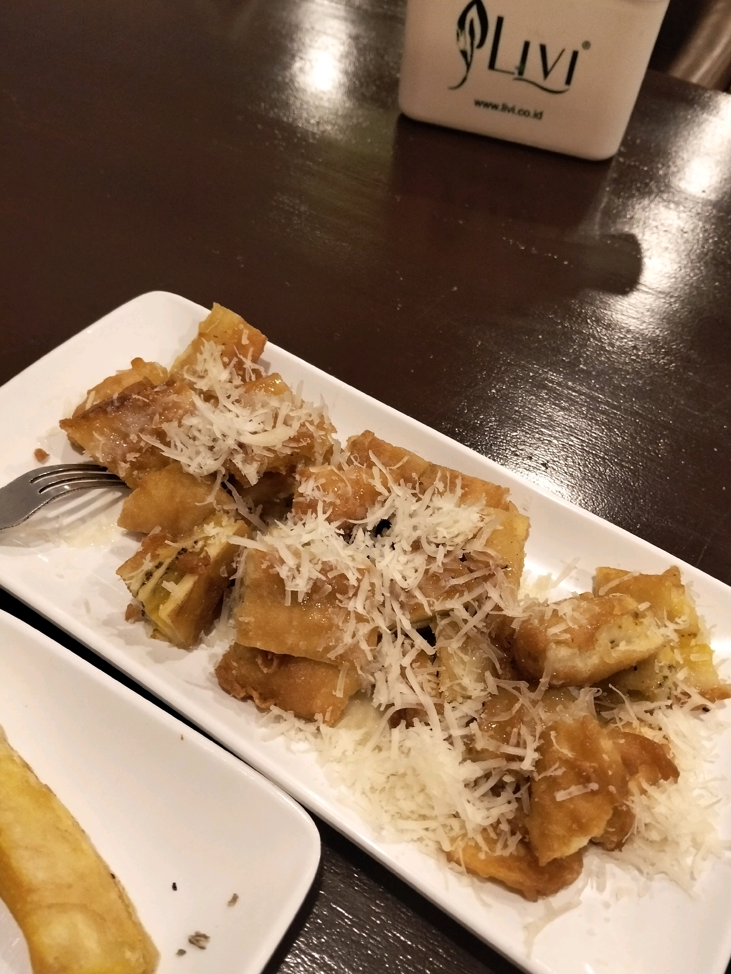 An overhead, close-up shot shows a white rectangular plate filled with fried banana pieces topped with grated cheese. A fork rests on the left side of the plate. The plate is set on a dark brown, reflective wooden table. In the upper right corner of the image, a white container with black text that reads "Livi" and "www.livi.co.id" is visible. The scene appears to be a food setting, possibly a restaurant. The location is Arang Limbung, Indonesia, suggesting this is a culinary scene from that region.
