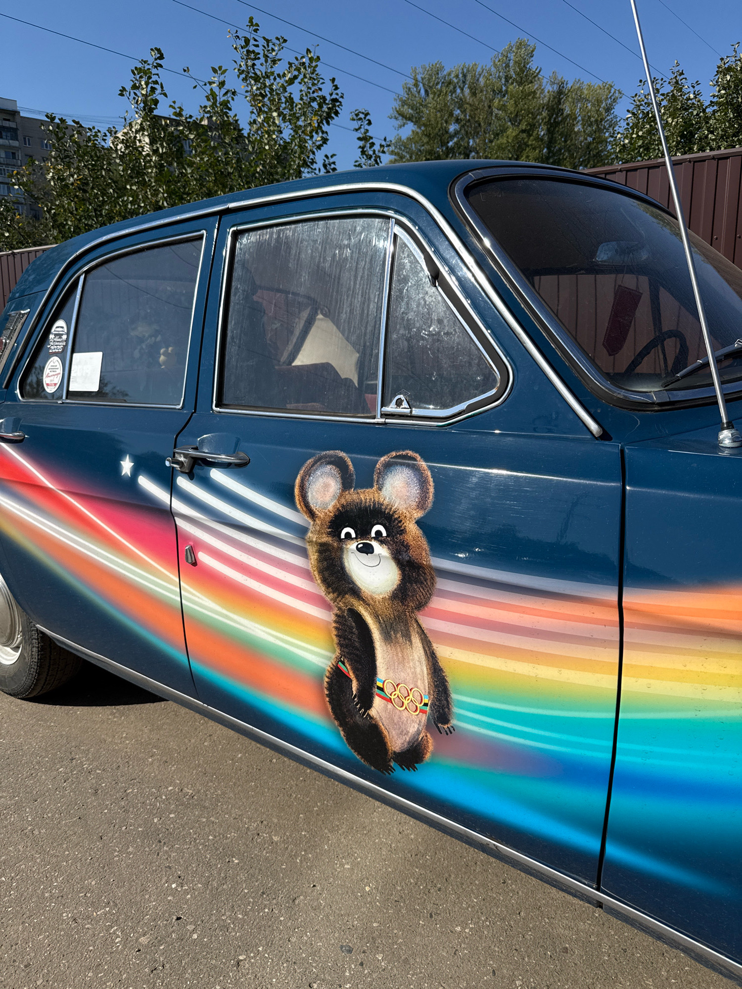 In Rusanovo, Russia, a vintage dark blue car is parked on an asphalt surface. The car features a distinctive graphic design on its side, depicting a rainbow with streaks of light and a cartoonish brown bear character wearing the Olympic rings. The bear has large ears, a friendly smile, and is positioned in the center of the graphic. A white star is also visible within the rainbow design, near the front of the car. The car's windows are tinted and reflect the surrounding environment, which includes trees and a brown fence. A metal antenna extends from the roof of the car. The sky is clear and blue.