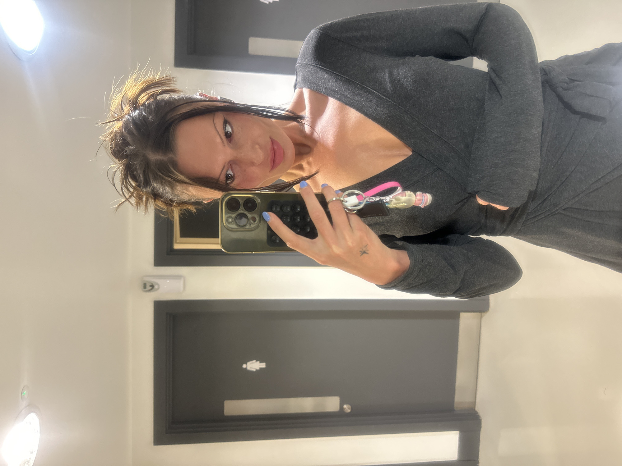 A woman is taking a selfie in a restroom. She is fair-skinned with brown hair and is wearing a gray robe or dress. She is holding a smartphone in her left hand and keys hanging from a pink lanyard are visible. A small tattoo is present on her wrist. The restroom has a modern design with dark gray doors and white walls. A female gender symbol is visible on one of the doors. The restroom features a round ceiling light. The location is unknown; there is no verifiable evidence indicating it is in London, United Kingdom.