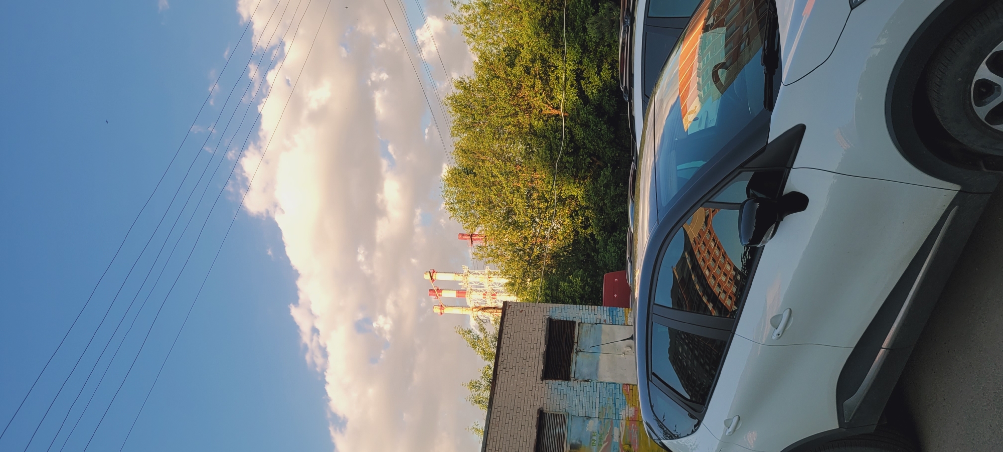 This image, captured in the city of Unknown, Unknown, presents a static outdoor scene under clear skies with scattered clouds. In the foreground, the left side of a white crossover SUV, featuring a black roof and black trim, is parked on an asphalt surface. Reflections of a multi-story building with a grid-like window pattern are visible in the car's side windows and windshield. Part of a second, lighter-colored vehicle is partially visible behind the first car on the right.
To the left in the midground, a single-story building constructed of light-colored bricks features two large, rectangular metal ventilation grilles above two closed, weathered metal doors. The lower facade of this building exhibits faded graffiti or mural elements in blue, green, and orange hues. A dense line of mature deciduous trees with vibrant green foliage forms a prominent barrier behind the vehicles and the brick structure, spanning across the midground. Several parallel power lines are visible horizontally across the upper midground and background.
In the background, above the treeline, stand multiple tall industrial chimneys or exhaust stacks. These structures are predominantly white with red bands or accents and display visible lattice-like support frameworks. The sky is bright blue with scattered, puffy cumulus clouds that are illuminated by warm, direct sunlight, suggesting late afternoon or early morning. A small, dark object, resembling a drone or distant aircraft, is faintly discernible in the upper central portion of the sky. No individuals are present in the depicted scene.