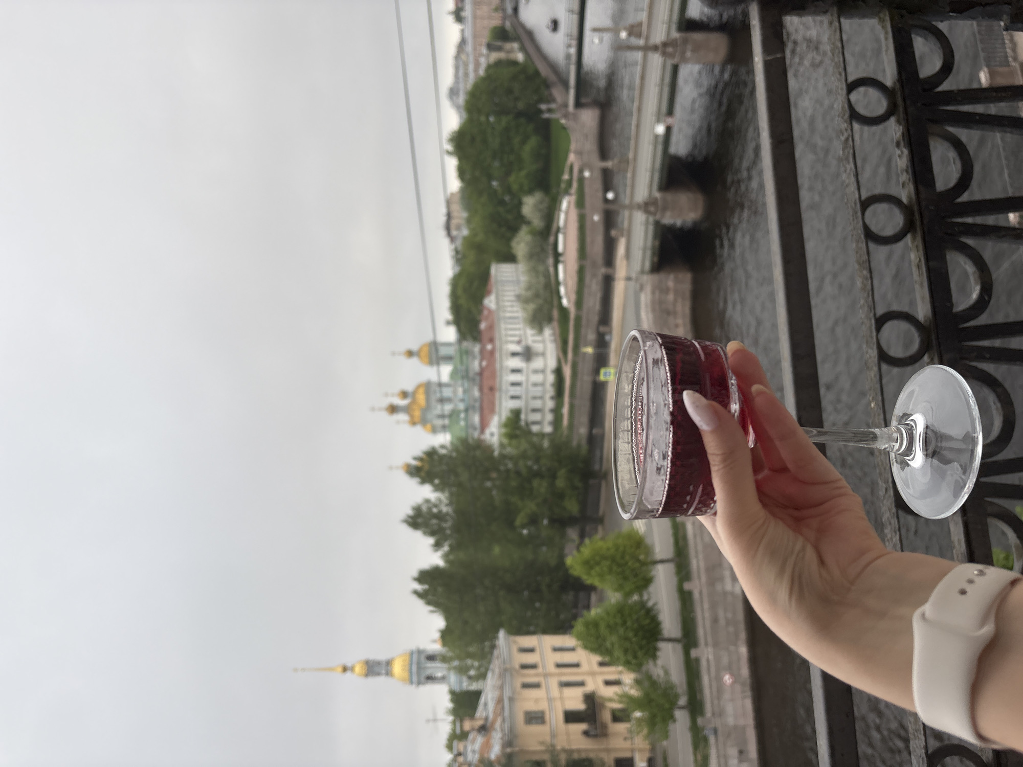 A horizontal, eye-level shot depicts a woman holding a wine glass filled with a red liquid, against the backdrop of Saint Petersburg, Russia. The woman's hand and arm are visible, wearing a white smartwatch, with manicured nails and pale skin. The wine glass is clear and filled with red wine.
The background is a cityscape, including buildings with golden domes and spires, typical of Russian Orthodox architecture. A river or canal flows through the city, with bridges visible in the distance. The sky is overcast and grey. In the lower right of the image is a black metal fence.