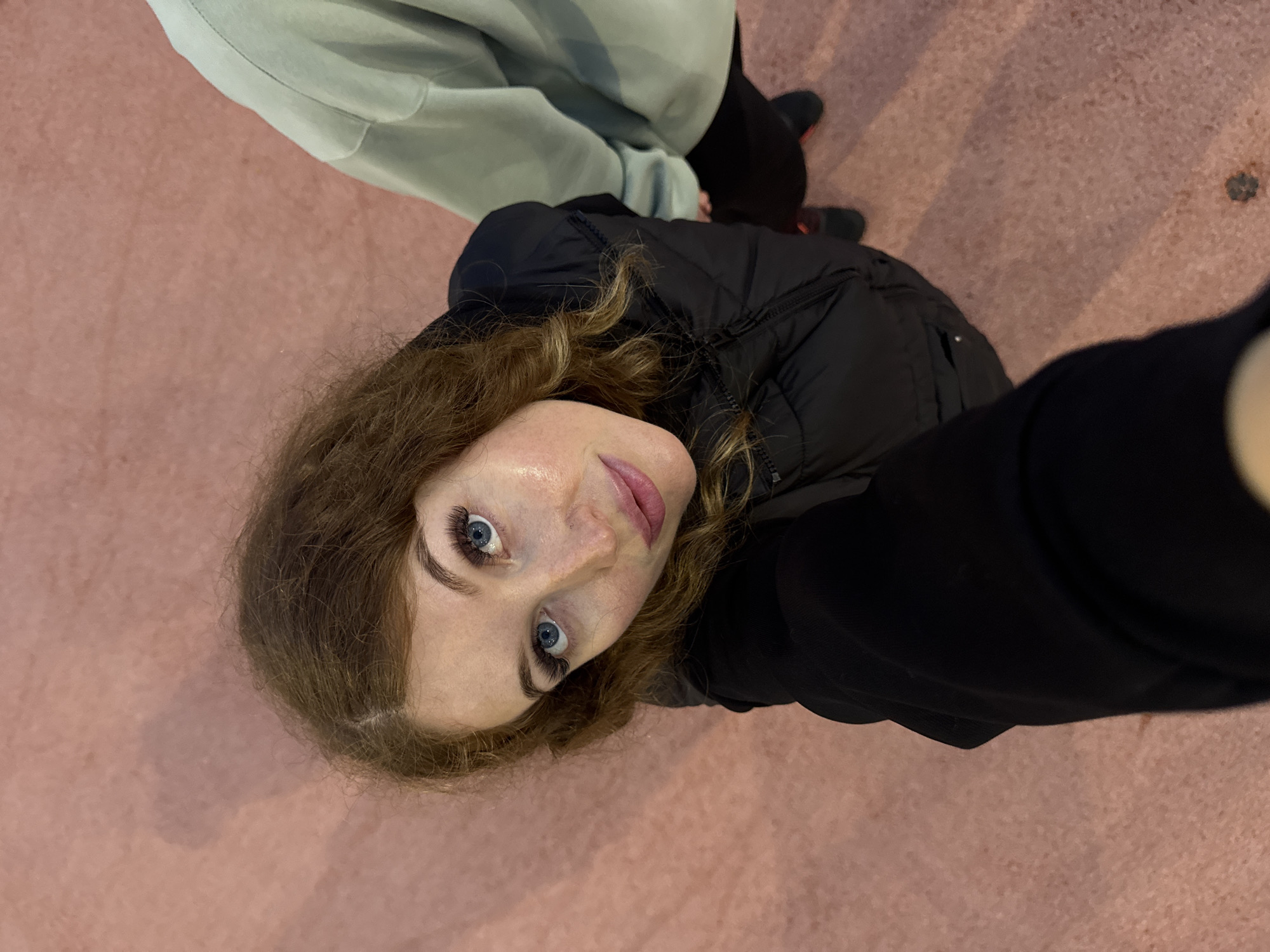 A medium-shot photograph, taken from a low, upward angle, depicts a young woman with light skin and shoulder-length wavy brown hair. She is wearing a black puffy jacket and looking directly into the camera with a slight smile. Her eyes are blue, and she is wearing makeup, including noticeable false eyelashes.
To the left of the woman, a person wearing light green sweatpants is partially visible. The floor appears to be a pinkish-tan concrete surface. The location is identified as Sirius, Russia.