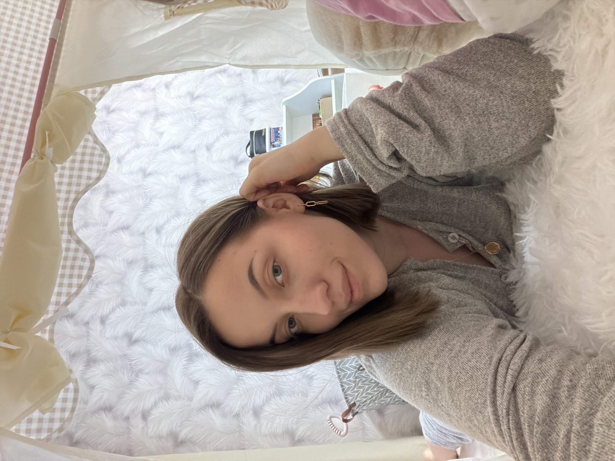 A woman with light brown hair is taking a selfie from inside a tent-like structure with beige and white fabric. The background features white walls with a subtle patterned design. A white shelf with items is visible behind her. The woman is wearing a gray sweater with gold buttons and a gold earring. The presence of a white furry object in the frame suggests she may be surrounded by soft furnishings. The location is unknown, with no objects to place the image in Reshetnikovo, Russia.
