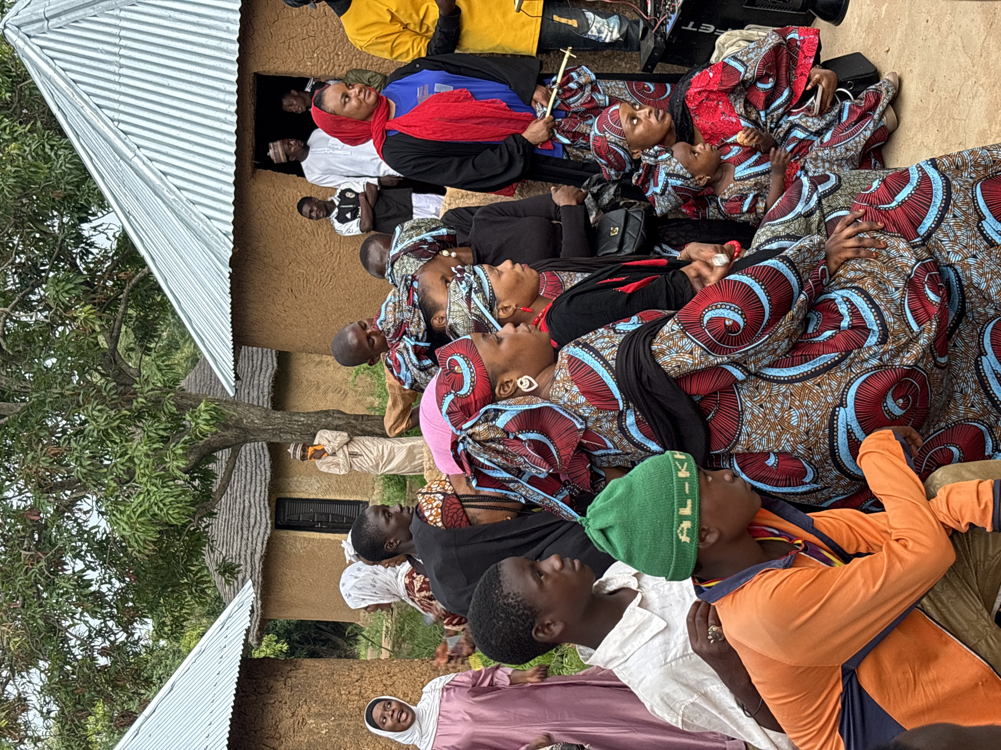 The image depicts a gathering of people, predominantly women and children, in an outdoor setting. The architecture suggests a rural or semi-rural location, with traditional mud buildings topped with thatched or corrugated iron roofs. The people are dressed in a variety of attire, with many women wearing colorful, patterned fabrics and headscarves. Some individuals appear to be engaged in conversation or observing the surroundings. The environment is characterized by trees and a dirt ground. The presence of modern electronic equipment, such as a speaker, indicates a potential event or activity taking place. The individuals are of various ages, and their expressions range from neutral to attentive. The scene appears to be a community gathering, possibly a celebration or a social event. Without further context, it is impossible to determine the exact location within the city of Jos, Nigeria, as the image does not contain any identifying landmarks.