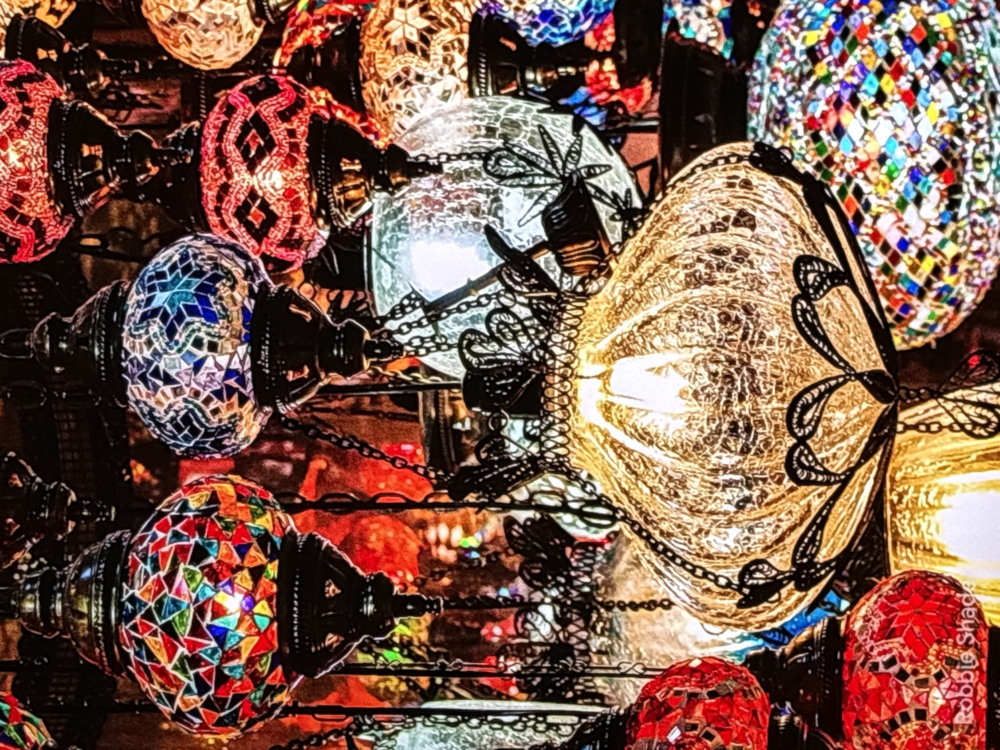 The image displays a close-up of several ornate, glass mosaic lamps, characteristic of traditional Turkish or Middle Eastern craftsmanship. These lamps are lit from within, casting a warm glow and highlighting the intricate patterns of colored glass. They are suspended by metal chains, creating a visually rich and textured composition. The background is dark, with hints of other colorful lamps and possibly a marketplace setting, suggesting an indoor display or vendor stall. The image does not contain any people, nor does it provide any specific visual cues that would allow for identification of the location as Krasnokamsk, Russia. The provided crops offer closer views of individual lamps, emphasizing their mosaic details and metallic hardware. The signature "Robbie Shade" is visible in the lower-left corner of the original image.