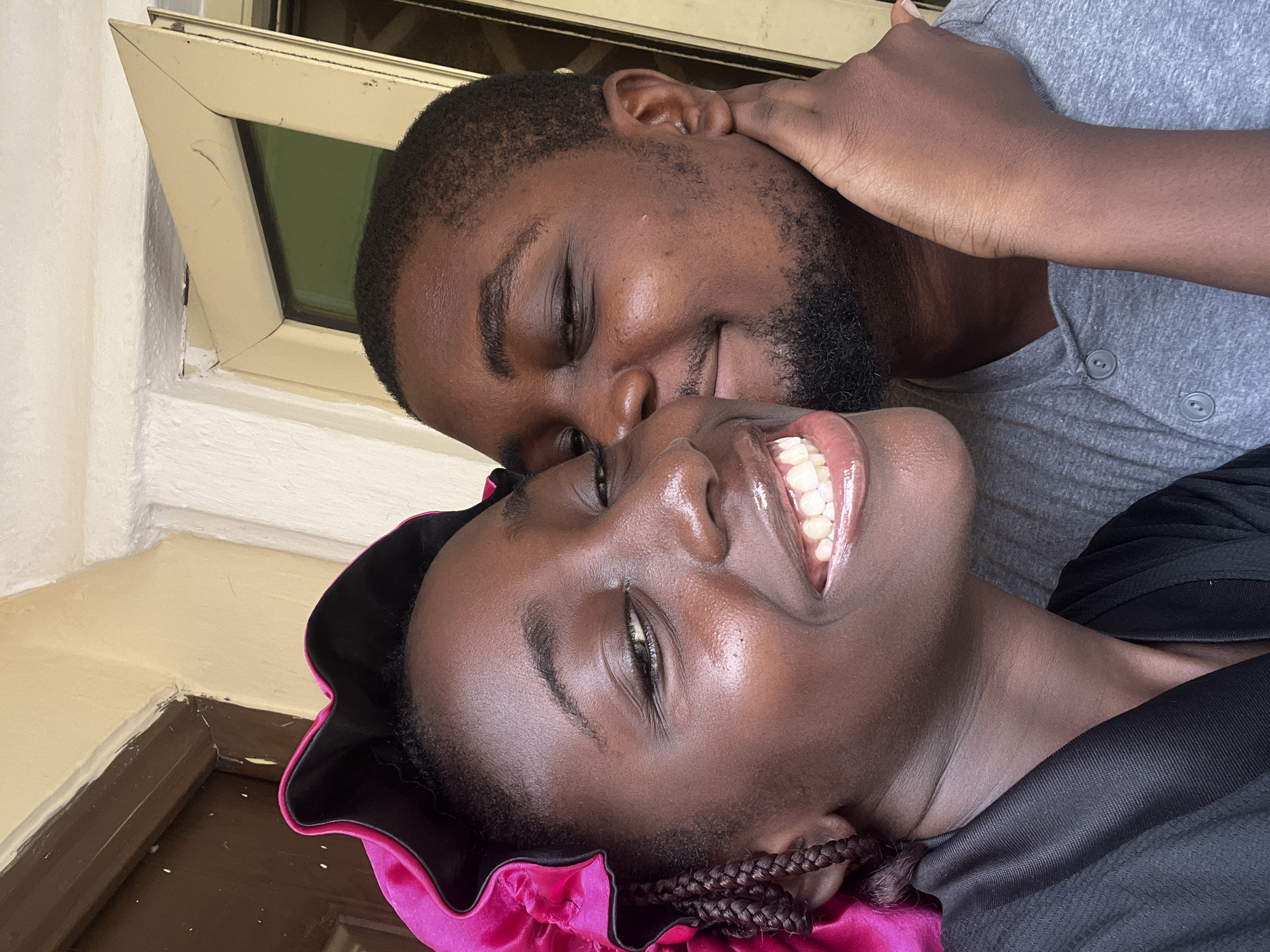 A close-up portrait of a young, dark-skinned Black woman and a Black man, likely in their 20s or 30s, shows them in an affectionate pose. The woman is smiling widely, revealing her teeth, while the man, with a beard and short hair, has his eyes closed and his arm around the woman's neck. The woman is wearing a dark-colored garment, partially obscured. She has braided hair and a bright pink or magenta satin bonnet. The man is wearing a gray shirt. The background includes a window frame. The location is presumed to be in the city of Abuja, Nigeria, based on external knowledge.