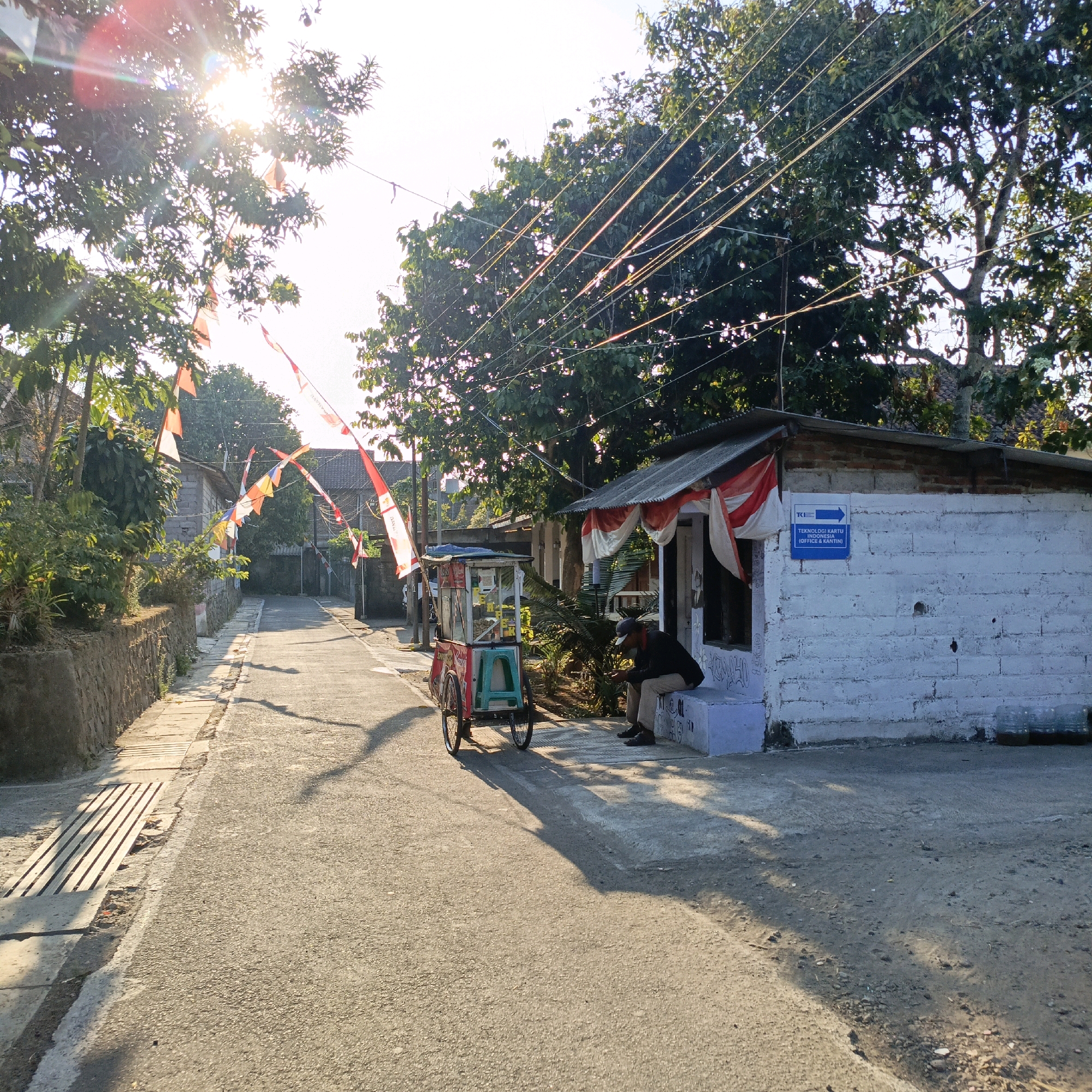 The image captures a sun-drenched, narrow street in Salatiga, Indonesia, likely in a residential or commercial district given the presence of a small brick building and a street-side vendor cart. The sun's rays pierce through lush green trees, creating bright lens flares and dappled shadows on the asphalt road. Red and white Indonesian flags are strung across the street, suggesting a festive occasion or national holiday.
On the right side of the street, a small, white brick building with a corrugated metal roof stands. It is adorned with red and white fabric draped over its entrance, reminiscent of the national flag. A blue sign on the wall indicates "TEKNOLOGI KARTU AKU" (Smart Card Technology) with an arrow pointing to the right, and a smaller text below it reads "(OFFICE & KANTIN)" (Office & Canteen). The building's wall has graffiti. A person in a dark jacket and light-colored pants is seated on a light blue bench outside the building, looking down at their phone.
To the left of the building, a colorful vendor cart with red and white branding is parked. It has a display case filled with items, and a light green stool is attached to its side. The cart has two spoked wheels.
The street itself is paved with asphalt, showing some wear and tear. A stone wall runs along the left side of the street, with a narrow sidewalk and a drainage grate. The overall scene depicts a typical street view in Indonesia, with vibrant colors, local commerce, and hints of national pride.