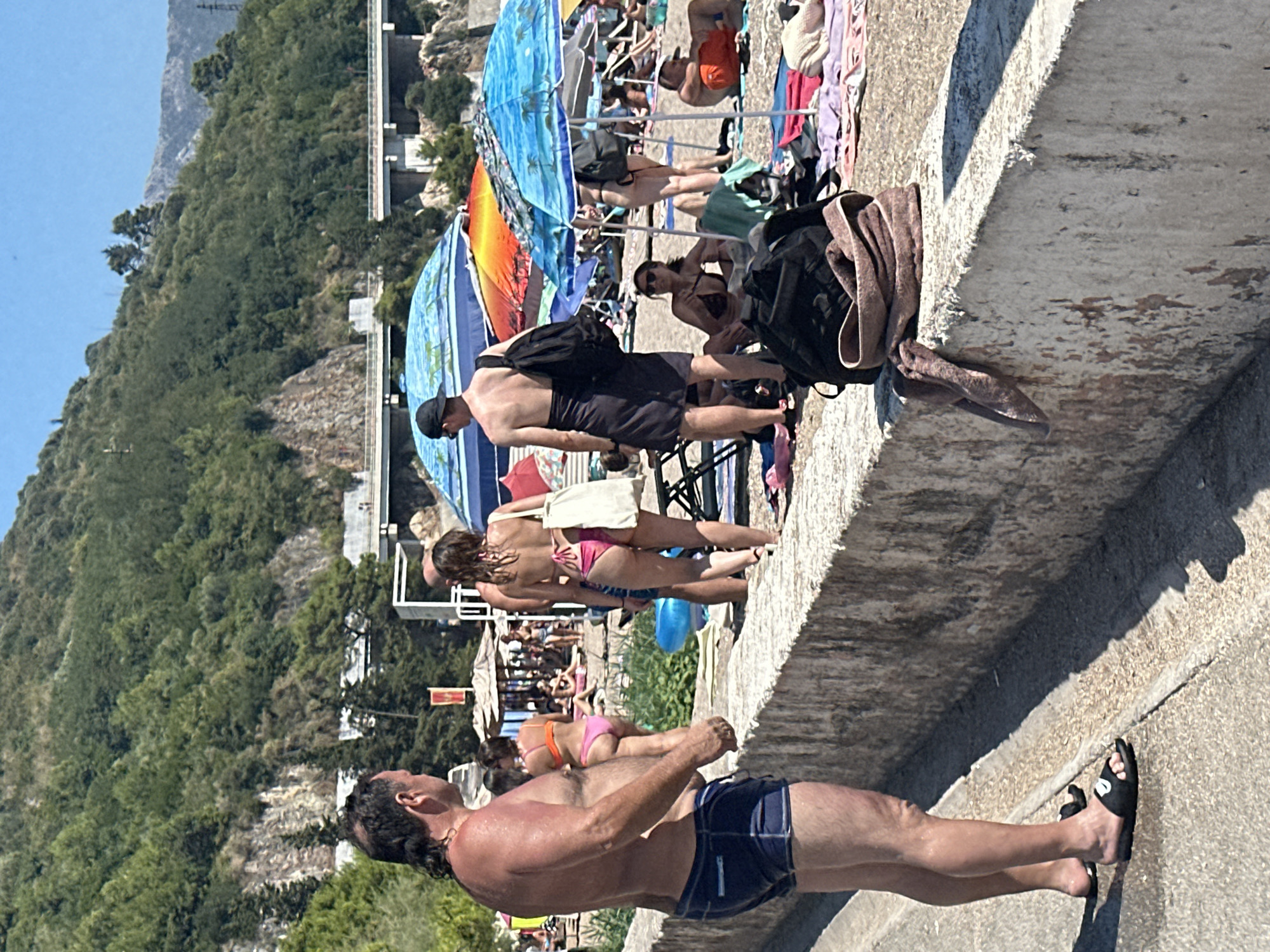 The image depicts a sunny day at a crowded beach in Budva, Montenegro. In the foreground, a man with a mullet hairstyle, wearing navy blue swim trunks and black slides, stands facing away from the camera, looking towards the beach. Behind him, several people are visible, including a woman in a pink bikini and a woman in a black bikini. Beach umbrellas in vibrant colors, predominantly blue and orange, provide shade. Beachgoers are seen sunbathing, sitting, and walking on the sand. The background features a steep, tree-covered hillside with a road or highway running along its base, and a clear blue sky overhead. The scene is characterized by the presence of numerous individuals enjoying a summer day at the seaside.