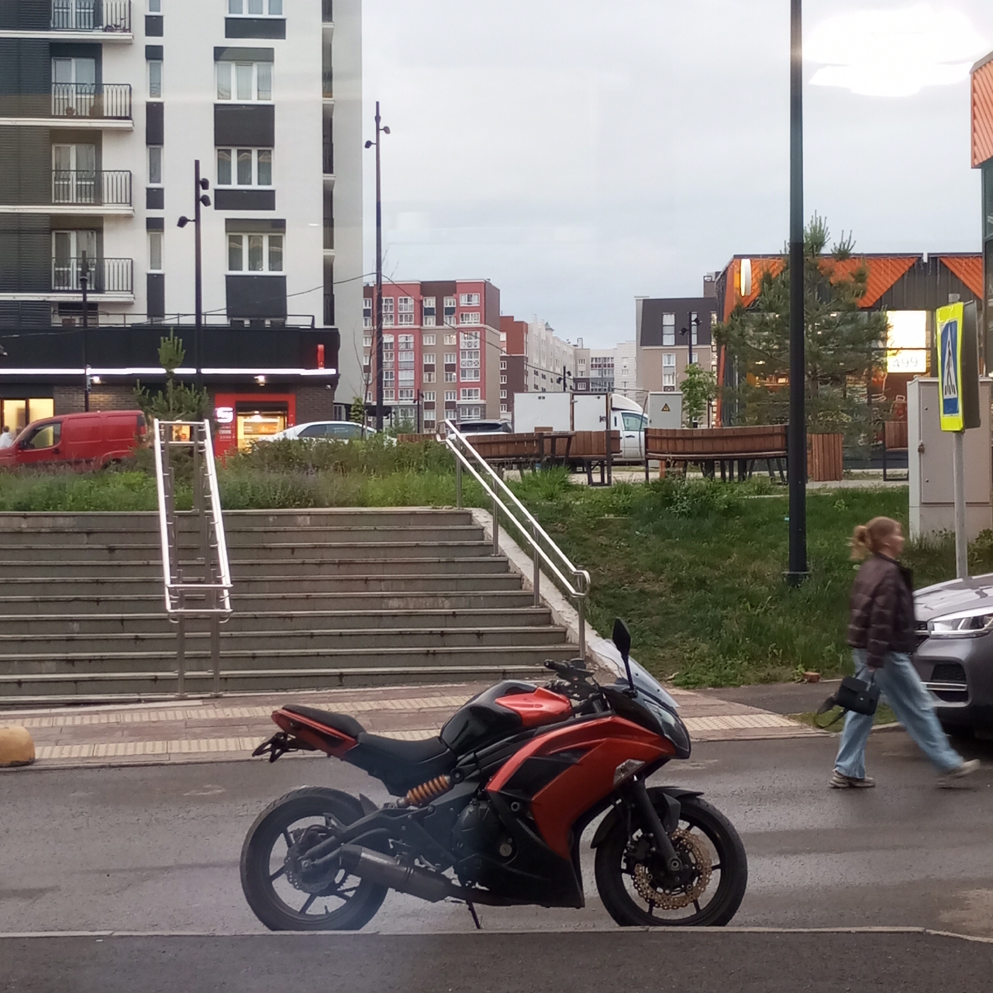 The scene depicts an urban environment in Pervomaysky, Russia, under an overcast sky. In the immediate foreground, an orange and black Kawasaki Ninja 650R motorcycle is parked on an asphalt street, facing right. The motorcycle features gold-colored front brake discs and a modified rear fender. To the left of the motorcycle's rear wheel, a yellow concrete bollard is visible on the sidewalk. A gray vehicle, possibly an SUV, is partially visible on the far right.
In the midground, a wide concrete staircase with multiple steps and two sets of metal handrails ascends towards the right. Above the stairs, a grassy embankment leads to a commercial area. A dark-fronted store with a prominent red "S" logo is visible, with a red delivery van parked directly in front of it on the left and a white car partially visible on the right. Across the street, a woman with blonde hair tied in a ponytail, dressed in a dark jacket, light blue jeans, and sneakers, walks from right to left, carrying a black shoulder bag. A yellow pedestrian crossing sign is located on the far right, near a utility box. Wooden benches and planters are situated in the grassy area on the right.
The background features several multi-story residential buildings. A white building with dark gray balconies and window frames is prominent on the left. In the center-background, a reddish-brown apartment building with numerous windows is visible. A tall, dark lamppost stands vertically in the center-right of the image. The ambient light indicates a cloudy day or late afternoon.