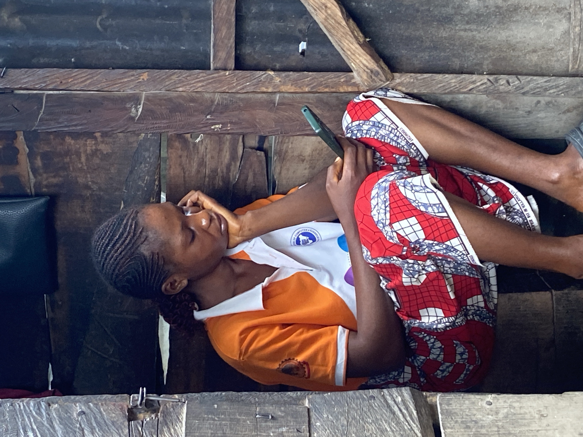 The image captures a young woman lying on her side, engrossed in her smartphone, within a rustic wooden structure in Mararaba, Nigeria. She has dark skin and her hair is styled in tightly braided cornrows. She wears a white and orange short-sleeved top; the white upper portion features a circular blue and white logo with partially visible text, "OF COMPUTER & SAMFORD...". An orange, gear-shaped logo is visible on her left sleeve. Her lower body is covered by a patterned wrapper or skirt in red, white, and blue geometric designs, with some visible text such as "CO0829T" and "SUPER WAX HOLLANDIS" on the fabric.
The woman holds a dark-colored smartphone in her right hand, her gaze directed towards its screen. She rests her head on her left arm, indicating a relaxed posture. The immediate surroundings consist of rough, unpainted wooden planks that form the surfaces and walls, suggesting an interior or sheltered space. The lighting appears to be natural daylight, indicating it is daytime. The overall scene depicts a moment of quiet personal engagement with technology in a simple, traditional setting.