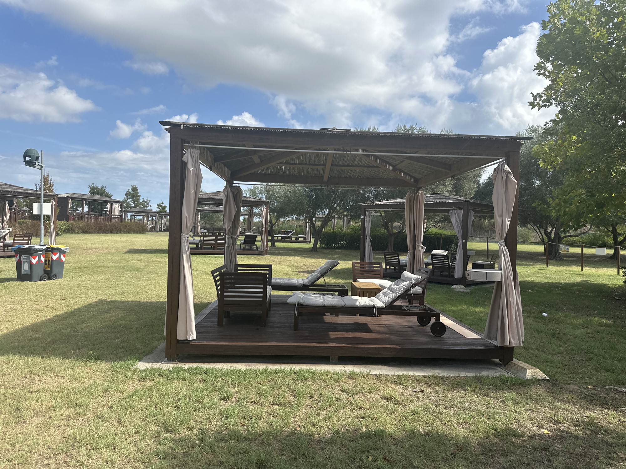 The image depicts an outdoor seating area with several covered gazebos, each furnished with lounge chairs and cushions. The gazebos are made of wood and have thatched roofs, with draped fabric on the sides. The area is covered in green grass, with some trees visible in the background. Two large trash bins are situated on the left side of the image. The sky is blue with scattered clouds. The scene suggests a leisure or resort setting. The style of the gazebos and the general ambiance are consistent with outdoor relaxation areas found in coastal or garden resorts, which are common in tourist destinations such as Ravenna, Italy.