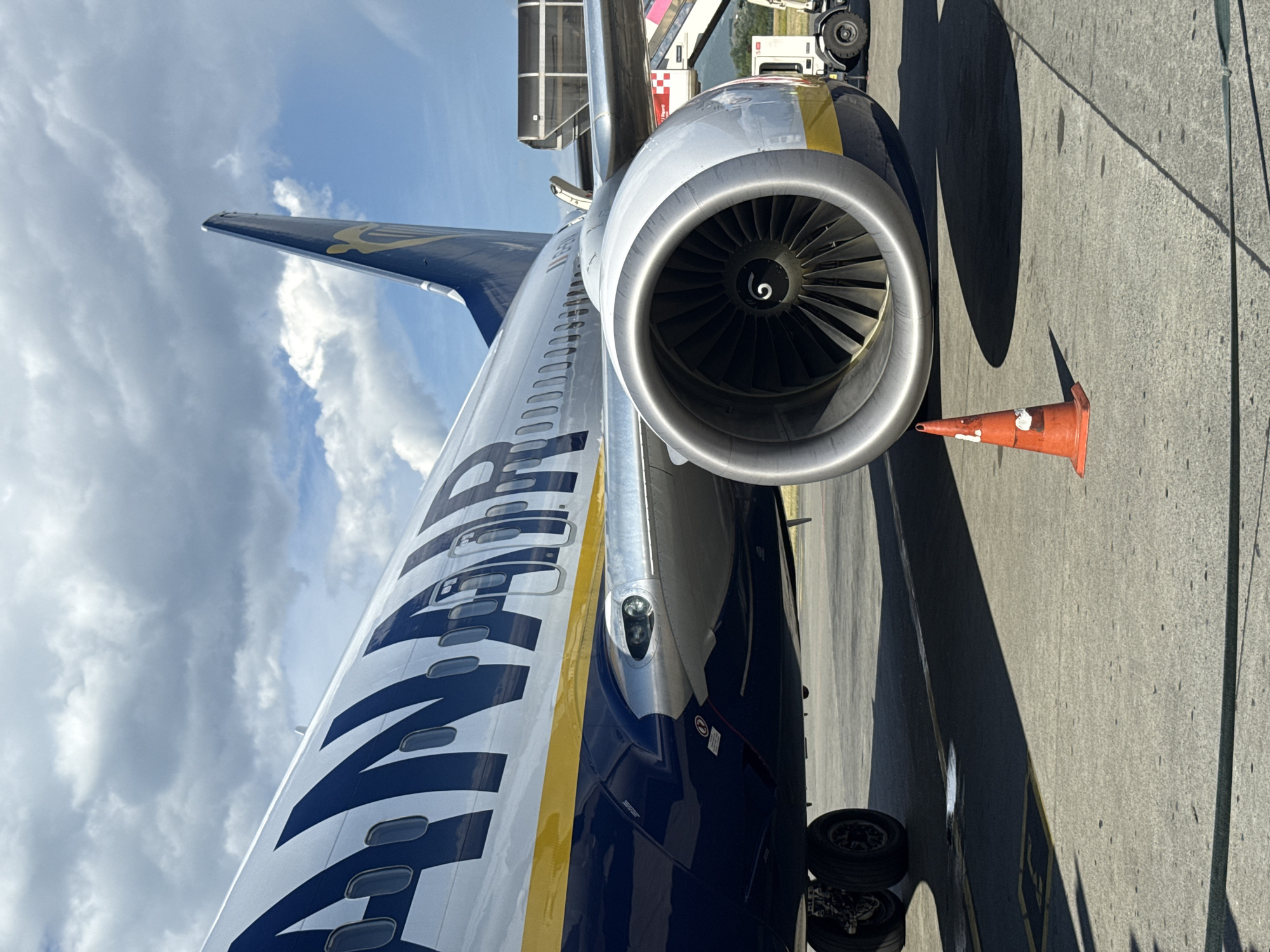A close-up, low-angle shot captures the engine and fuselage of a Ryanair airplane parked on a tarmac under a partly cloudy sky. The airplane's livery features large navy blue lettering that spells out "RYANAIR" against a white background, with a distinct yellow stripe running along the fuselage. The massive jet engine, made of polished metal, dominates the foreground, its fan blades visible within the intake. A single orange traffic cone with white markings stands in the foreground, casting a shadow. In the background, a portion of an airport jet bridge or staircase and a white service vehicle are visible. The scene is set on a concrete surface marked with yellow lines and other subtle pavement details. The city of Kruja, Albania, is not discernible from the image content, nor are any people or specific actions depicted.