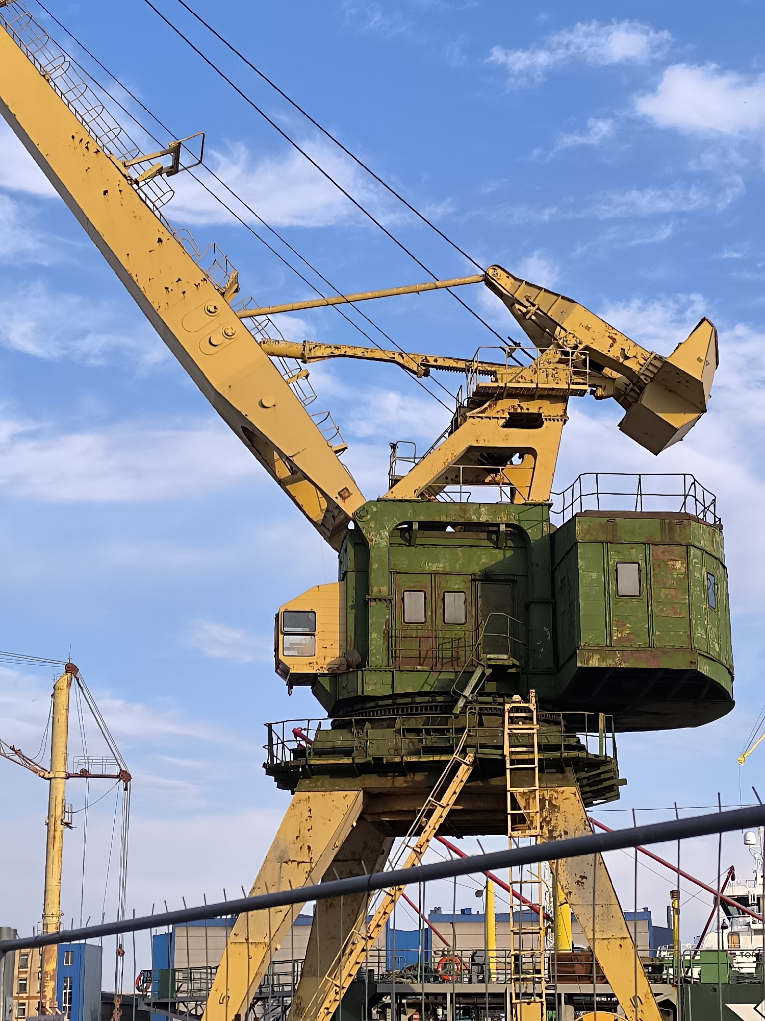 This is a close-up, eye-level, vertical, medium shot of a large, yellow and green industrial crane against a blue sky with scattered white clouds. The crane is weathered, with visible rust and peeling paint, suggesting it is an older piece of equipment. It is positioned in a port or industrial area, likely in Klaipėda, Lithuania, given the context of other cranes and structures visible in the background.
The main subject is the upper portion of the crane. The long, yellow boom extends diagonally from the upper left towards the center of the frame. It is supported by a substantial green housing unit. This housing unit appears to be a cabin or control room, with visible windows and an access ladder leading up to a platform with railings. There are also visible cables and wires running across the sky, connecting to the crane and potentially other machinery.
In the background, parts of other industrial structures and cranes are visible, including another yellow crane to the left and elements of ships or port buildings. A black metal fence runs across the lower portion of the frame, partially obscuring the view of the base of the crane and the ground. No people are clearly discernible in the image, though a small red object on a platform might indicate a person. The lighting suggests it is daytime, possibly late afternoon or early morning, given the angle of the light and the shadows. The overall impression is one of industrial activity and maritime infrastructure.