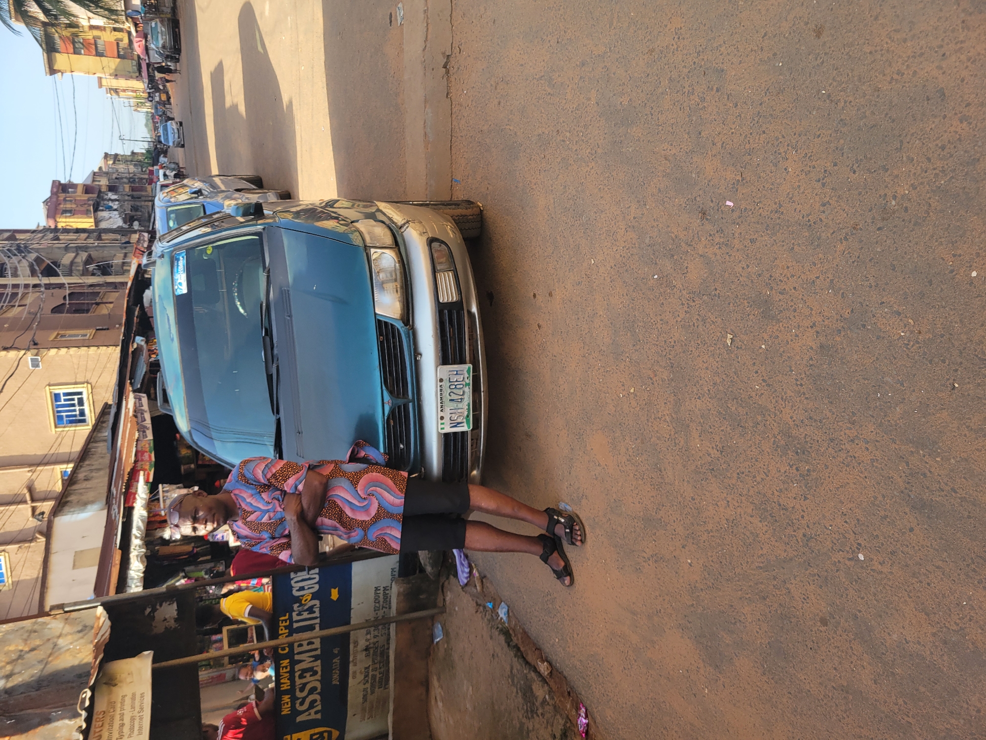 In Onitsha, Nigeria, on a sunlit street, a man in a colorful patterned shirt and black shorts stands with his arms crossed, leaning against a teal minivan. The minivan, parked on the right side of the road, has a license plate reading "ANAMBRA NSH 428EH". Adjacent to the minivan, on the left, is a stall with a sign for "NEW HAVEN CHAPEL ASSEMBLIES OF GOD". Various goods are visible in the stall, and other people can be seen in the background. The street is paved with reddish-brown asphalt, and buildings with multiple stories line the right side of the road, indicating an urban environment. The overall scene depicts a street-level view of a commercial area in Onitsha.