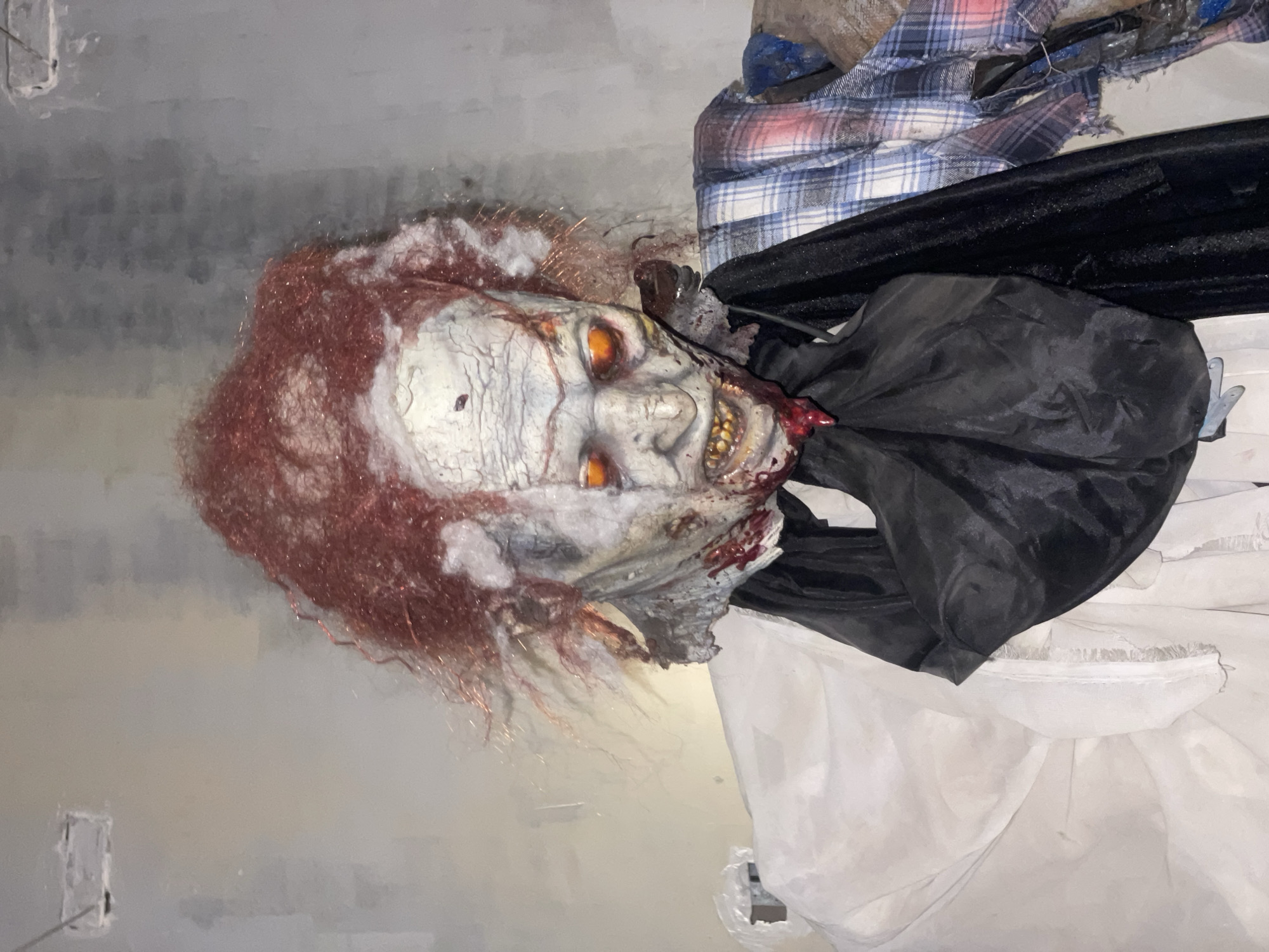 A Halloween prop depicting a decaying, zombie-like clown is shown. The prop has patchy red hair, glowing orange eyes, and a grotesque, decaying face with exposed teeth and bloodstains. It is dressed in a white shirt, a black scarf, and a tattered plaid shirt. The background appears to be a wall, possibly in a storage area or a room being decorated for Halloween. No specific location in Denpasar, Indonesia, can be determined from the image.
