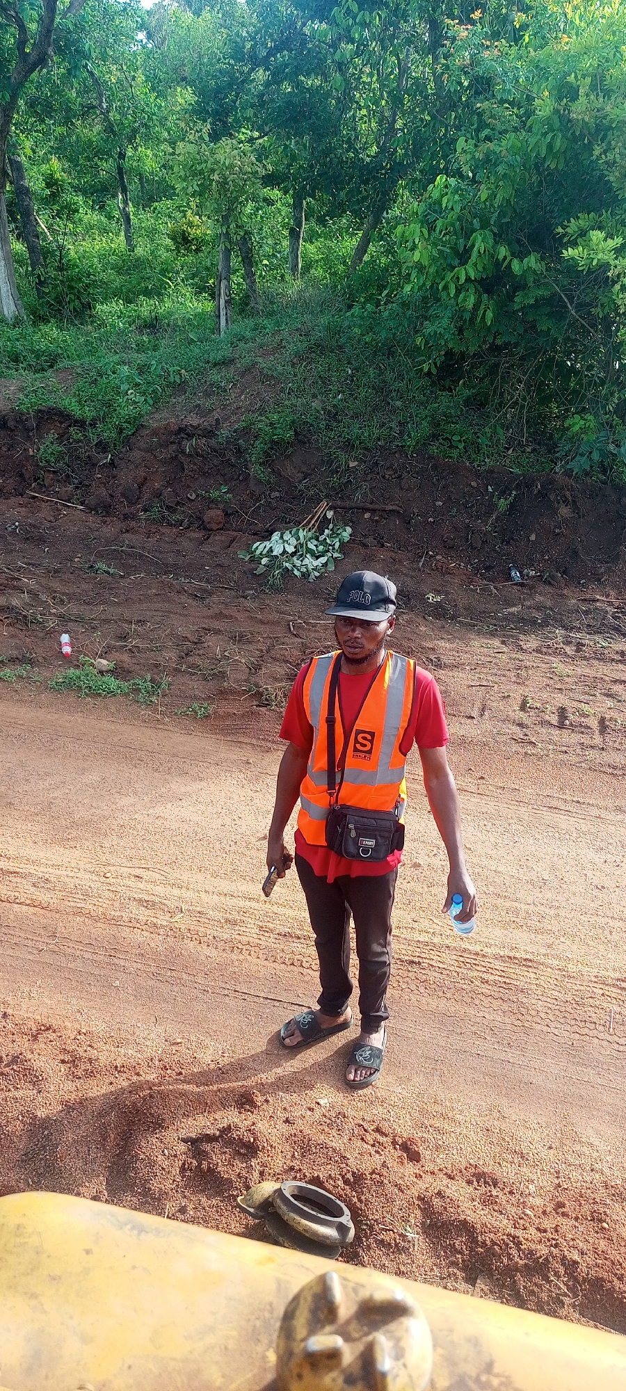 A man stands centrally on a dirt road, facing slightly to his left. He is wearing a red t-shirt, black pants, and dark slides/sandals. Over his t-shirt, he sports an orange safety vest with reflective strips; a large white "S" and the word "SMALEYE" are visible on the chest. A black baseball cap with "POLO" written on it covers his head, and a black shoulder bag hangs across his body. In his left hand, he holds a dark object resembling a smartphone, and in his right hand, a clear plastic water bottle.
The setting is outdoors, characterized by an untarred, reddish-brown road. To the man's left, there's a slightly raised bank of earth with sparse green growth and some discarded plant material. Further in the background, a dense cluster of green trees and bushes forms a natural backdrop, suggesting a rural or undeveloped area. A red plastic bottle and another clear bottle are visible as litter on the bank. In the immediate foreground, the top of a yellow piece of machinery is partially visible, hinting at construction or heavy equipment activity.
The time of day appears to be daytime, with bright, natural light illuminating the scene. The man's expression is neutral.