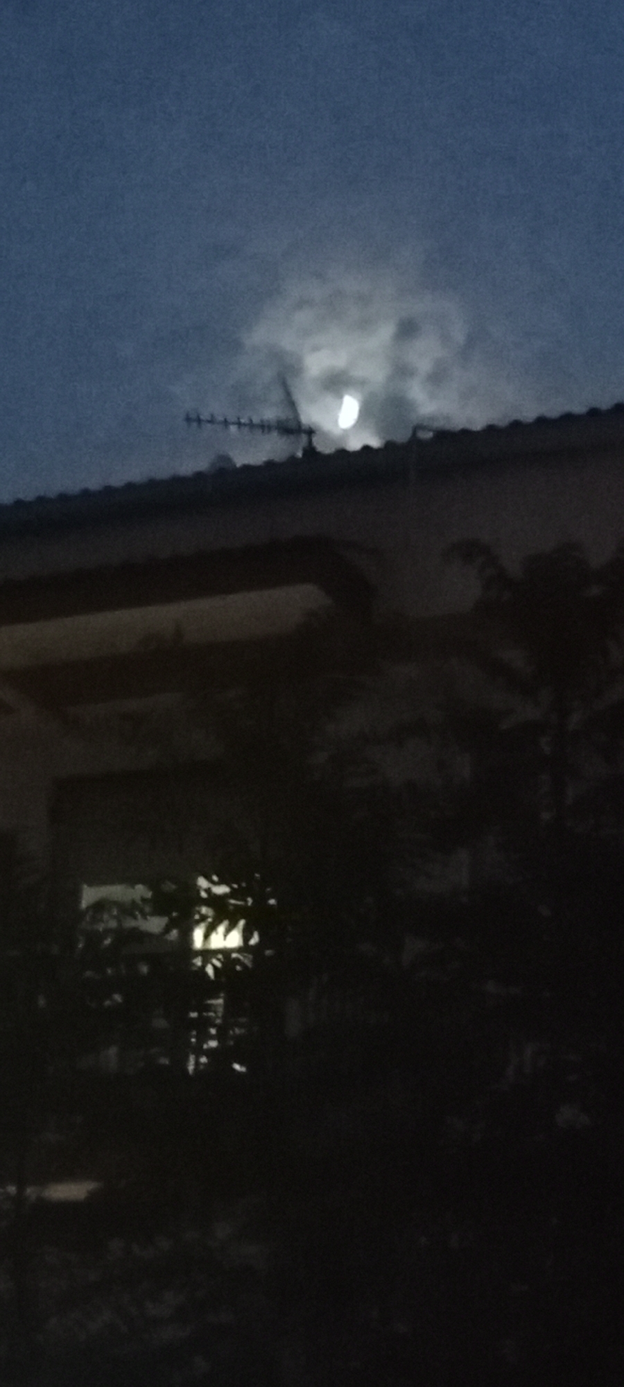 The image depicts a nighttime scene with a partially obscured moon and some clouds in a dark blue sky. In the foreground, a dark silhouette of a tiled roof extends across the frame. A television antenna is visible on top of the roof. Below the roofline, the scene is very dark, with the shapes of trees and foliage suggested. A faint light emanates from a window in a building in the background, illuminating some of the surrounding vegetation. The overall impression is of a quiet residential area at night in a location that might be near Galapagar, Spain.