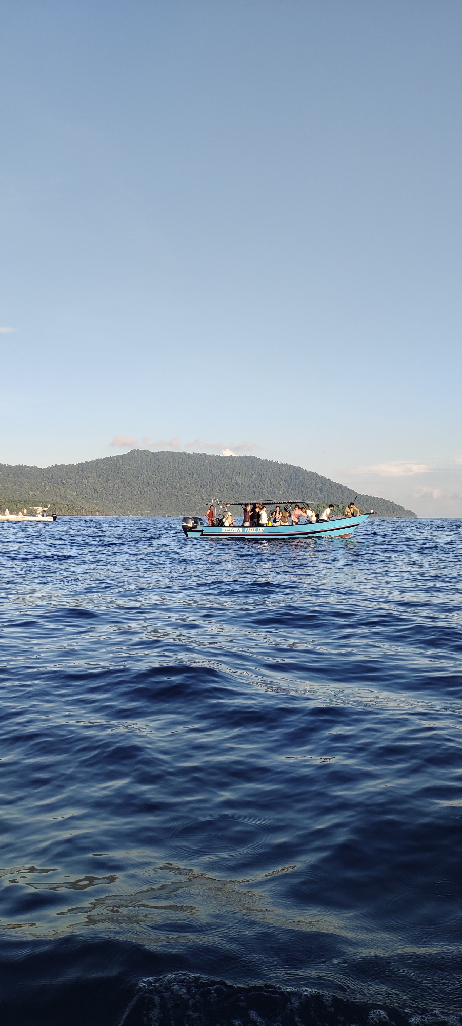 A group of people are on a boat in the ocean near a jungle-covered island. The boat has "SCUBA HOLIC" written on its side. The water is blue with small waves. The sky is clear and light blue. The island has a green, tree-covered mountain. Another, smaller boat is in the distance to the left. The scene appears to be in the city of Sabang, Indonesia, likely a tourist excursion.