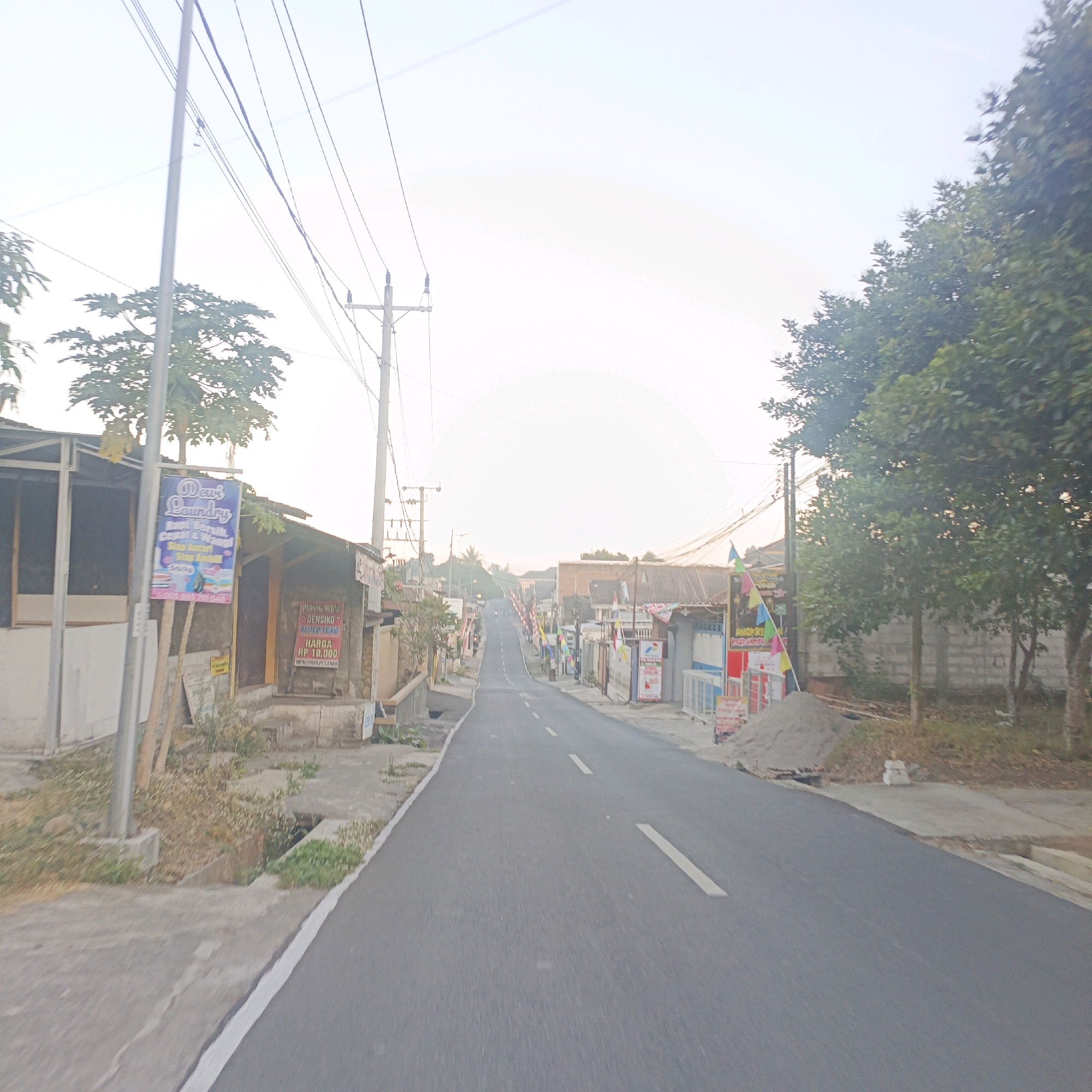 The image depicts a street scene in Salatiga, Indonesia, characterized by a paved road with white dashed lines, leading uphill and curving slightly to the left. The road is flanked by buildings and vegetation. On the left side of the road, a building with an open front structure features a sign for "Dewi Laundry." Further up the street on both sides, various small shops and houses are visible, some adorned with flags and colorful banners. A pile of sand is present on the right side of the road. Overhead, numerous power lines stretch across the sky, supported by utility poles. The overall atmosphere suggests a residential or commercial area in a tropical climate, with the bright, hazy sky indicating either early morning or late afternoon light. There are no people clearly visible in the frame.